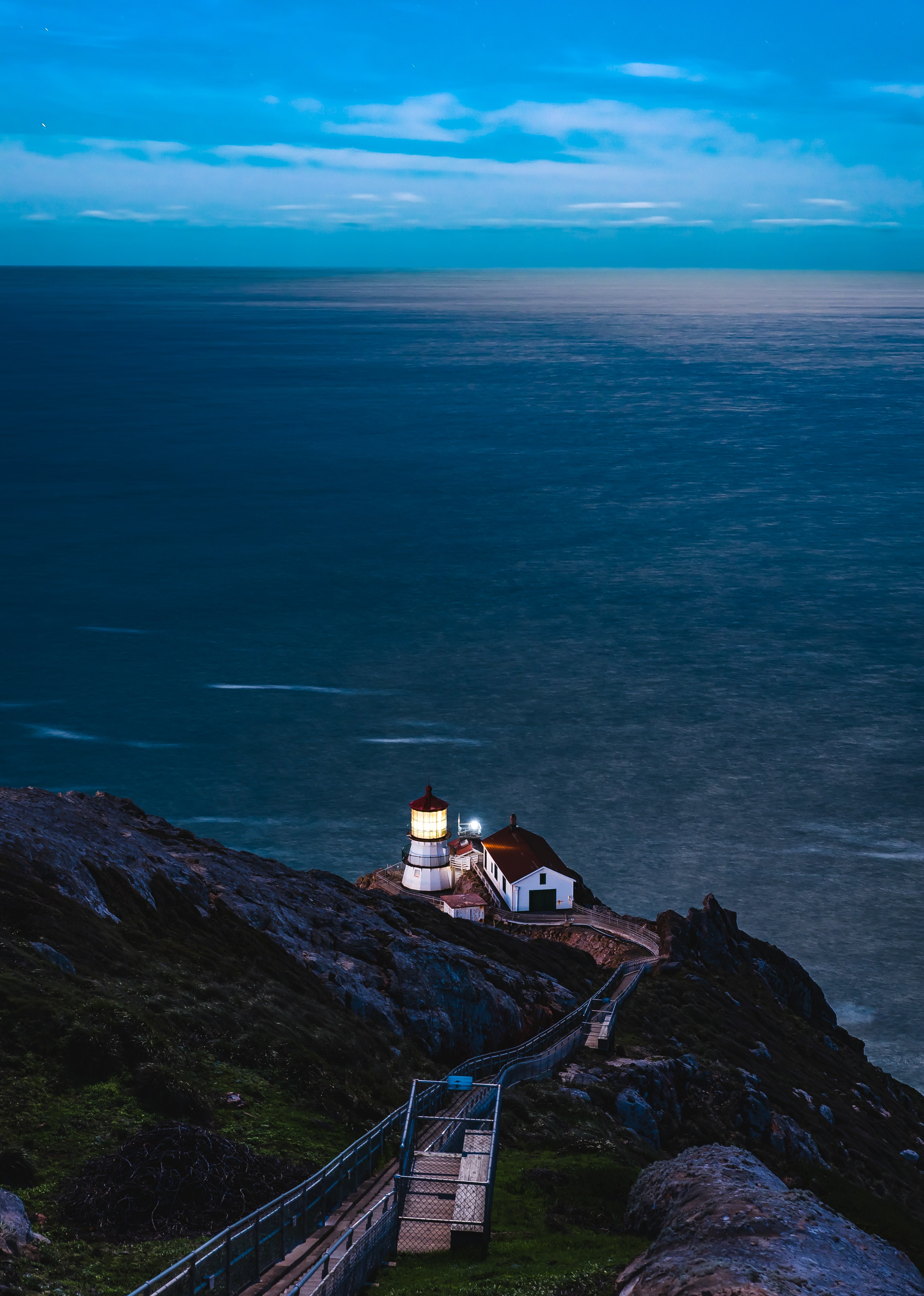 white and red light house with a lit light and barn next to it on a cliff overlooking the sea, night light 4k, Phone HD Wallpaper