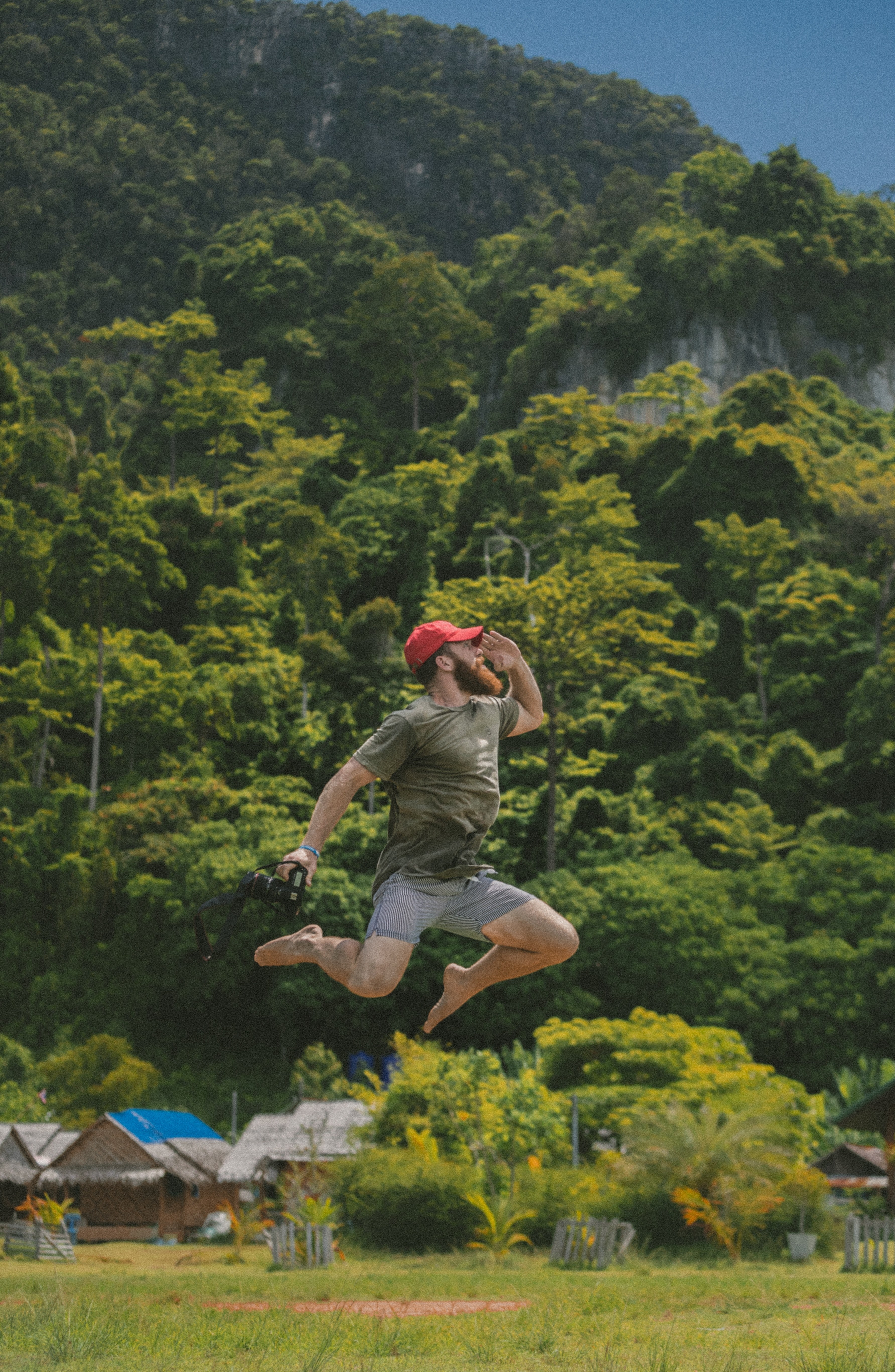a bearded man in a red baseball cap holding a camera jumps before green hills, floating in phi phi 4k, Phone HD Wallpaper