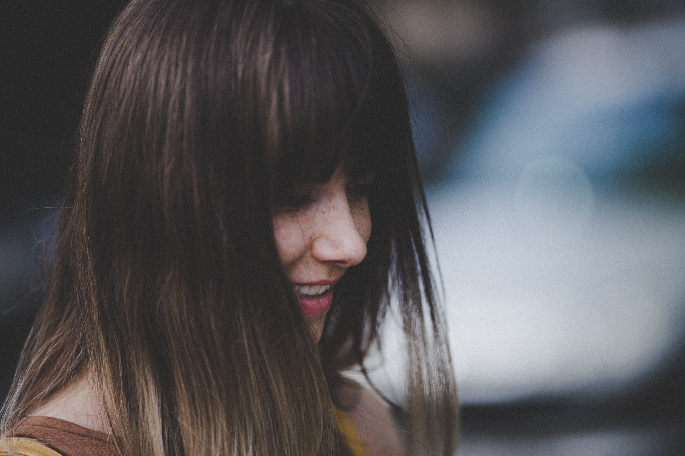 a woman with long brown hair and bangs smiles while looking downward against a blurry background, woman with bangs smiles 4k, HD Wallpaper