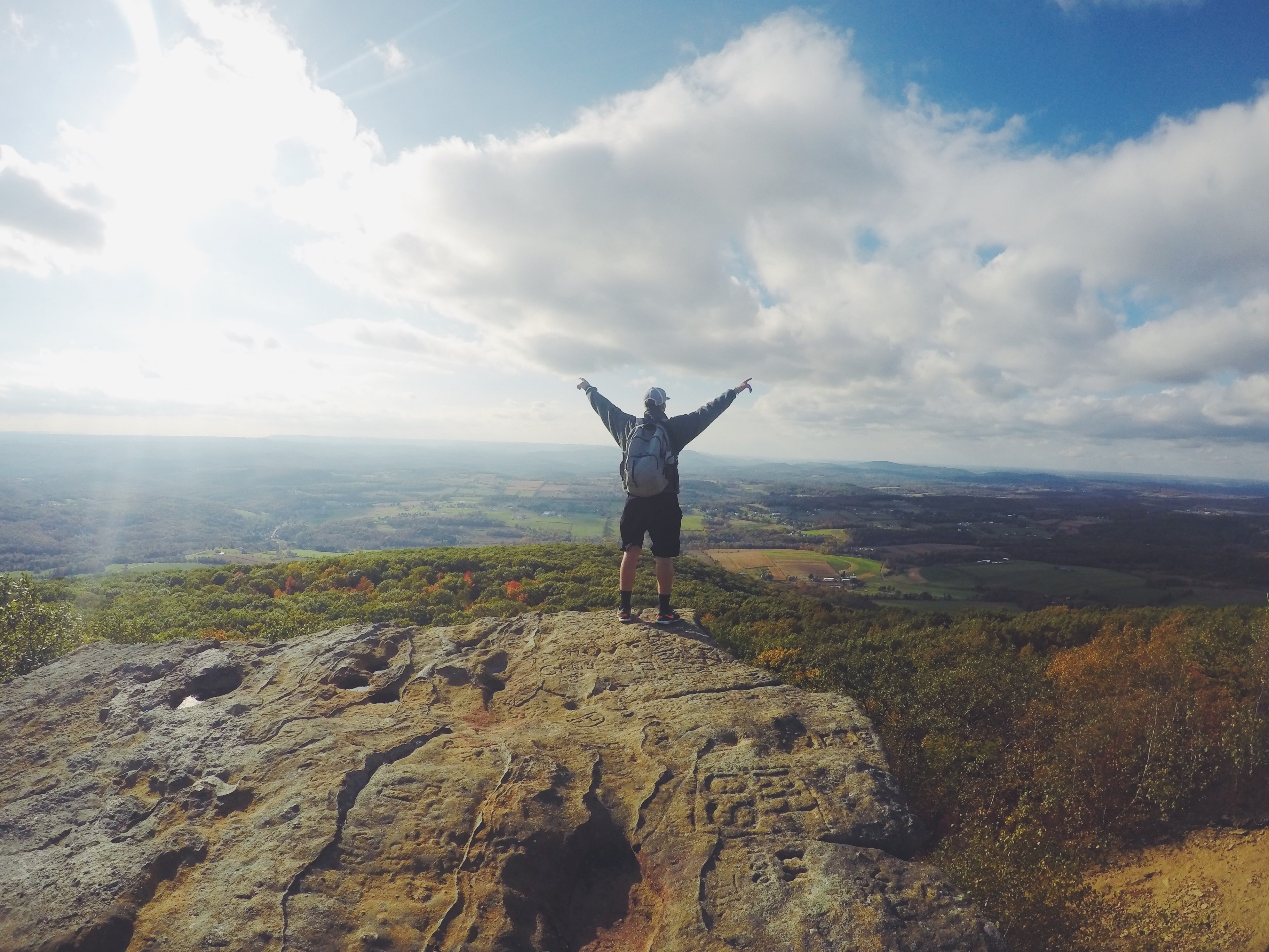 a man with a backpack standing with his hands outstretched on a rocky ledge overlooking green countryside, high above the green plains 4k, HD Wallpaper