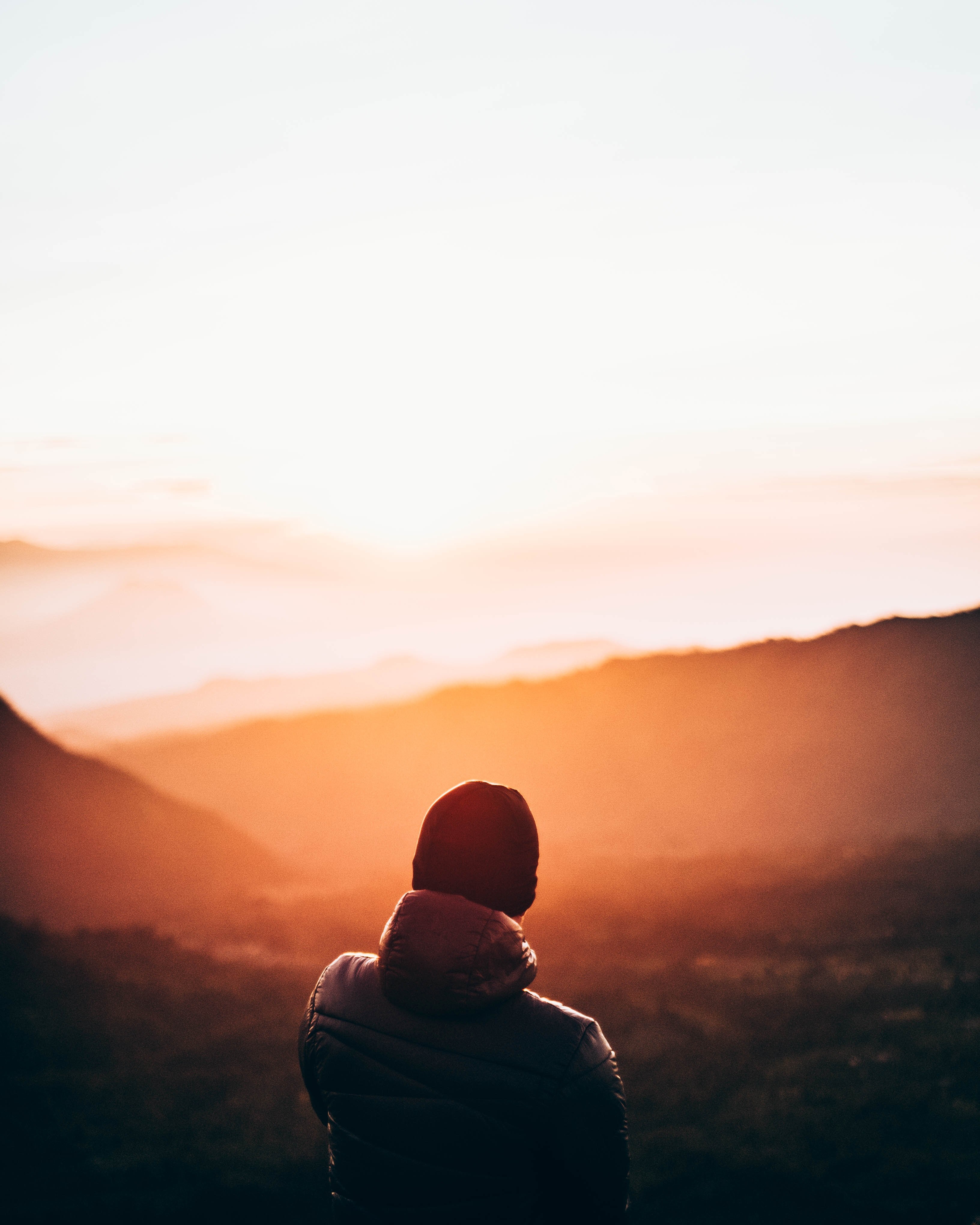 a person wearing a hat and hooded jacket looking at the sun over distant mountains, mountain sunlight sunset and sunrise hd, 4k, Phone HD Wallpaper