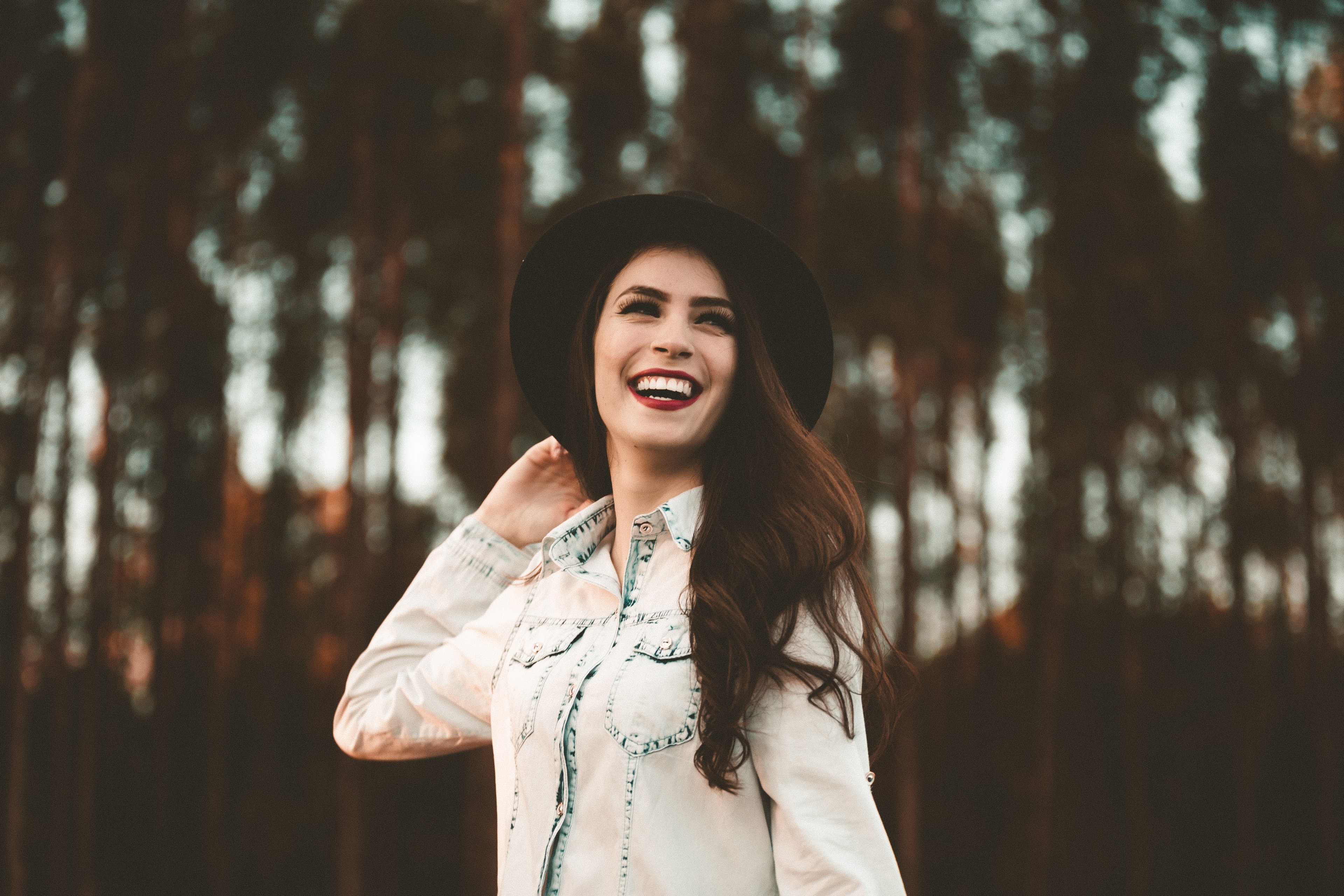 a woman in dark lipstick and a black hat smiles broadly in front of tall trees, woman in hat with tall trees 4k, HD Wallpaper