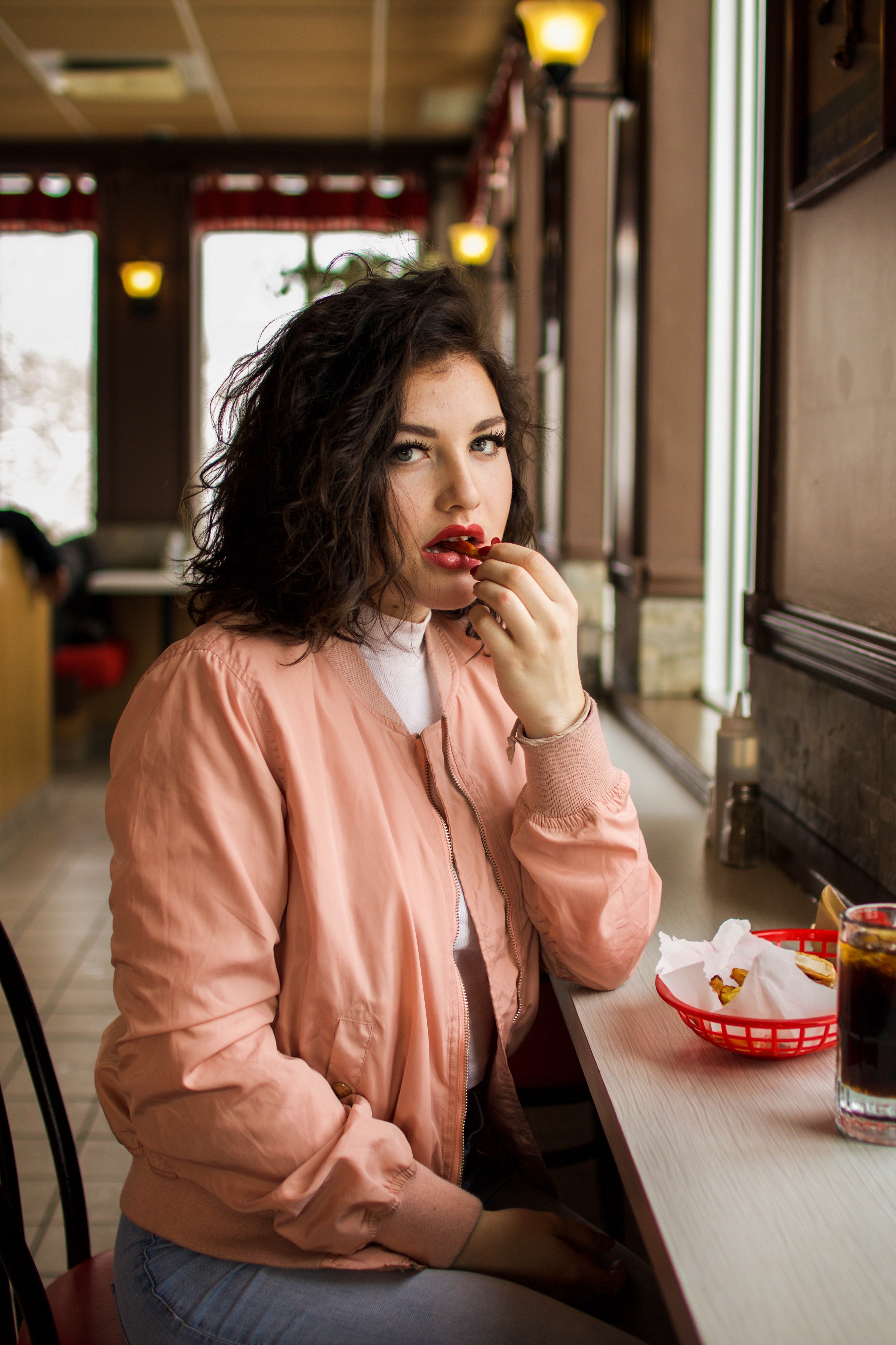 a woman in a pink jacket sits at a counter eating and looking at the camera, woman eating at counter 4k, Phone HD Wallpaper