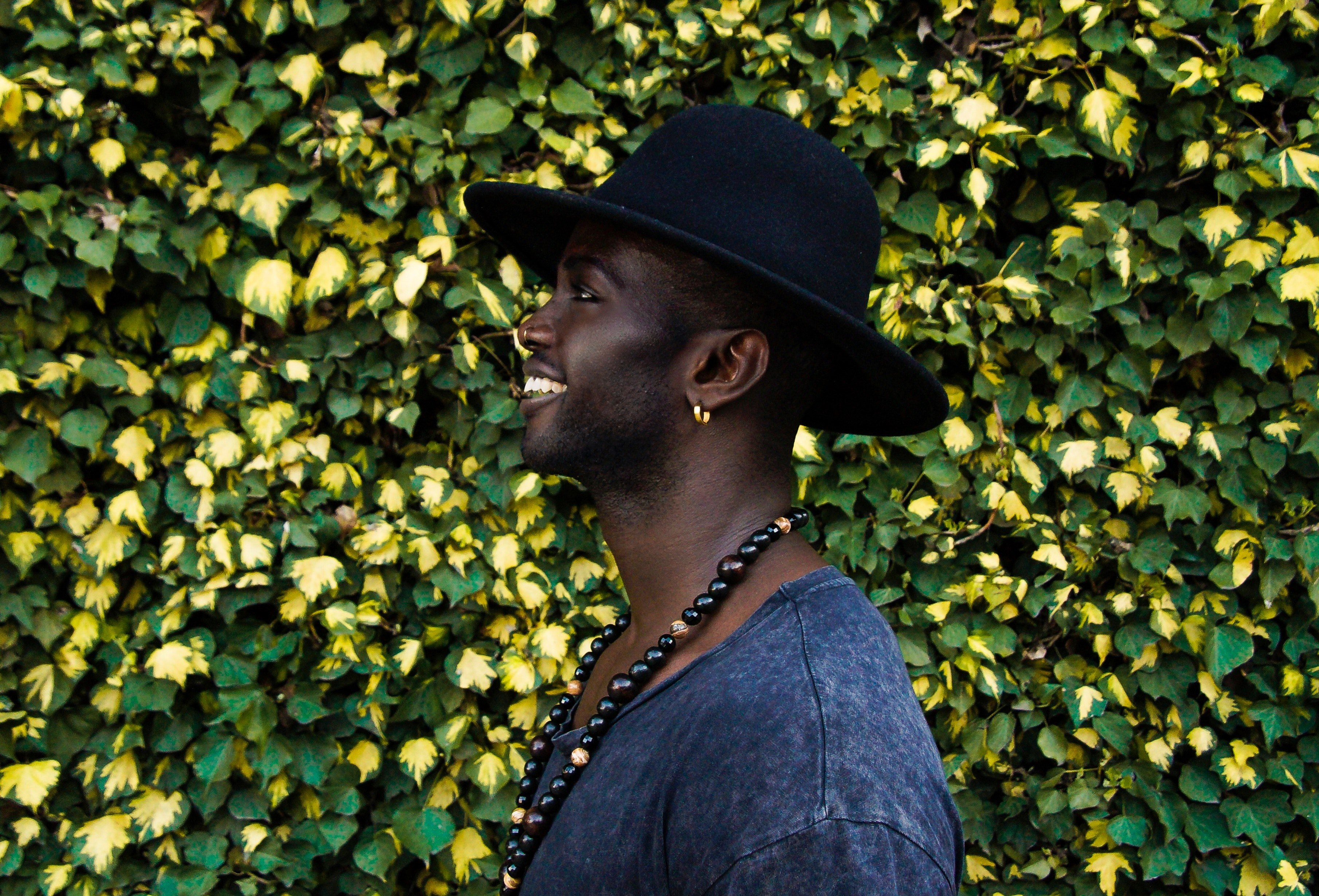 man in a hat and necklace in front of a tall hedge smiling to the side, forever muse 4k, HD Wallpaper