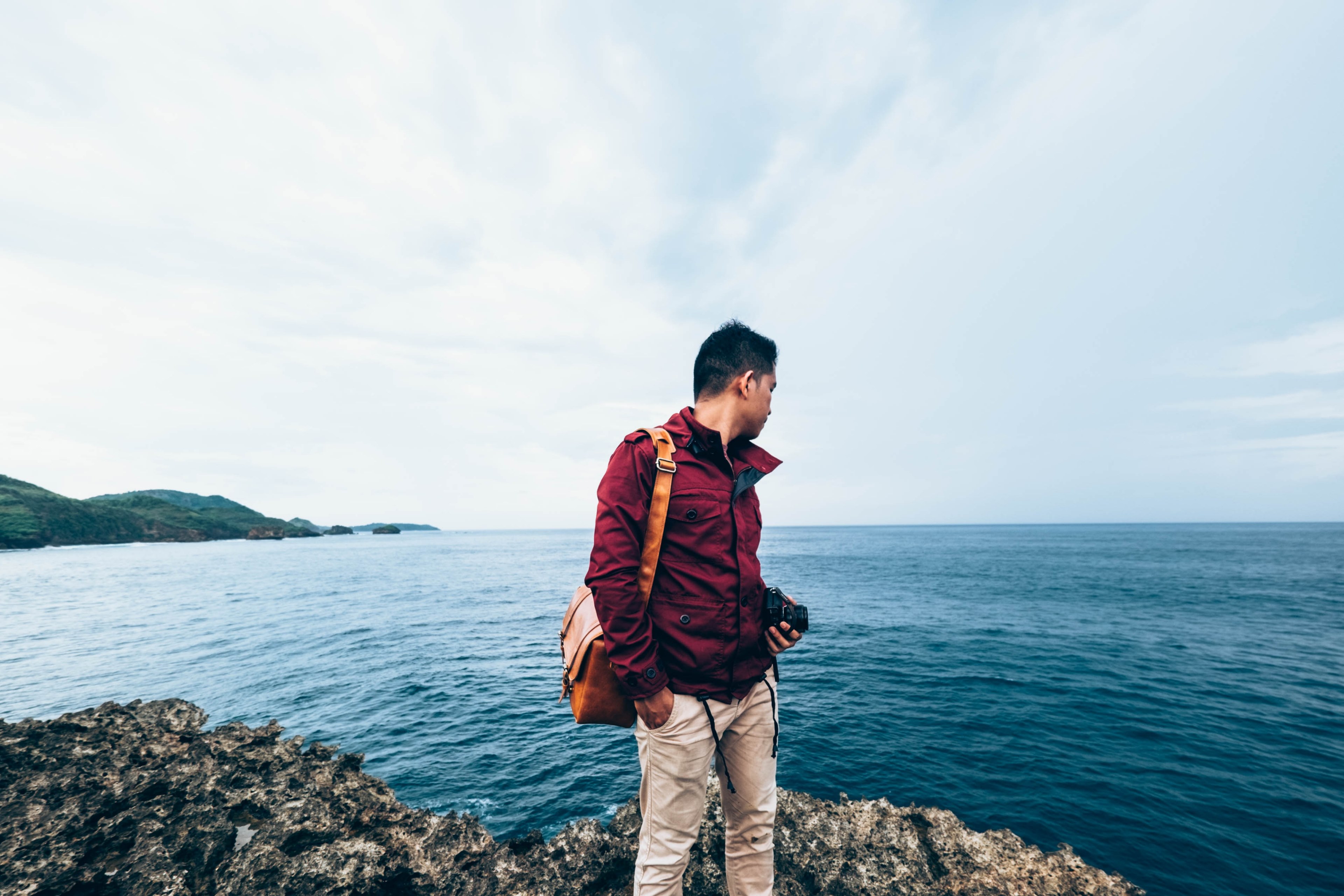 a man wearing a red jacket holds a camera and bag by the coast, man by the coast 4k, HD Wallpaper