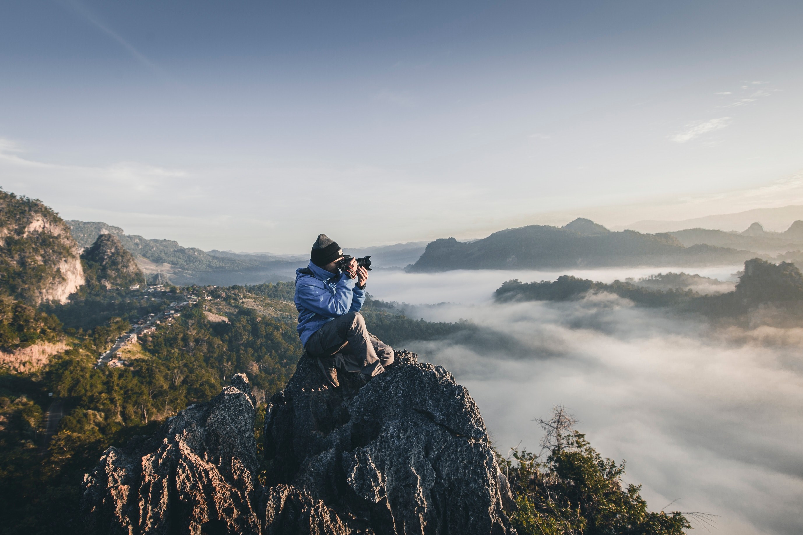 a man with a long lens camera sitting on top of jagged rocks overlooking wooded hills, above the clouds around the mist 4k, HD Wallpaper