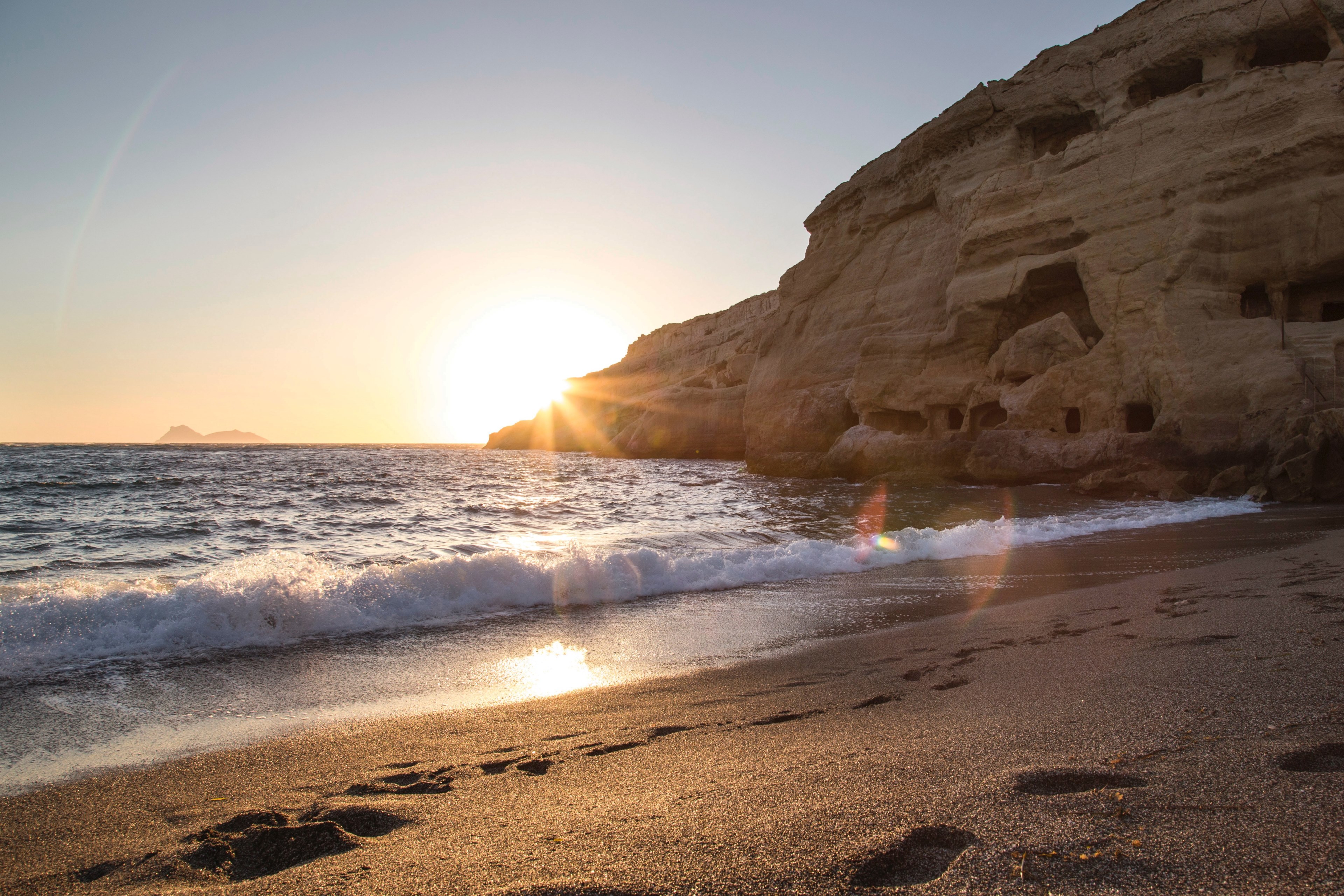 a course sand beach in matala with foot marks along the shoreline leading toward a large rock coastline_ small waves crash into shore and the bright sunrise or sunset creates a yellow glare 4k, HD Wallpaper