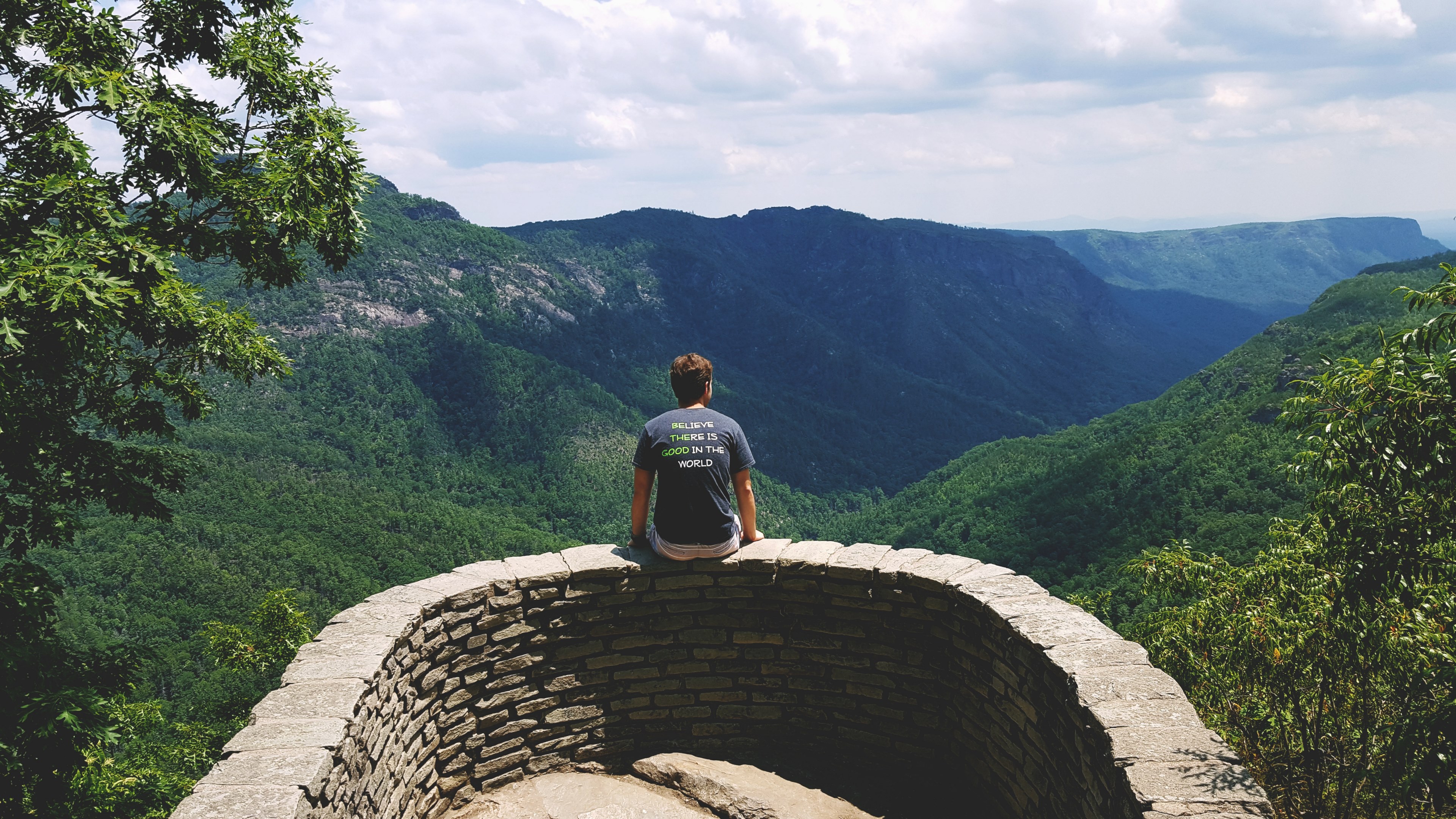 a man sitting on a small stone wall overlooking a green mountain valley, small wall by a mountain valley 4k, HD Wallpaper