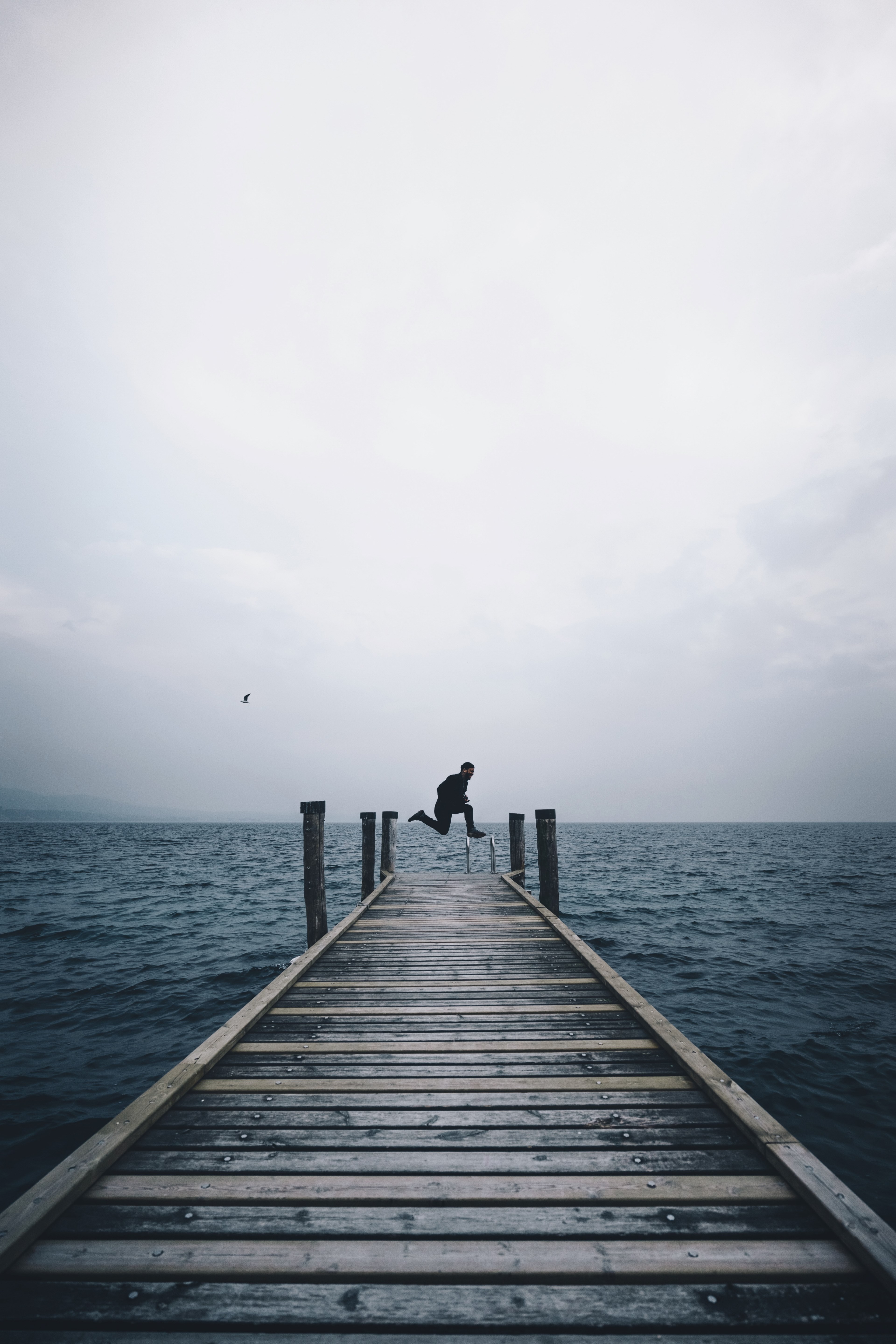 a person jumping in the air on the edge of a dock by a lake in punta saint vigilio on a cloudy day, punta saint vigilio dock jumps 4k, Phone HD Wallpaper