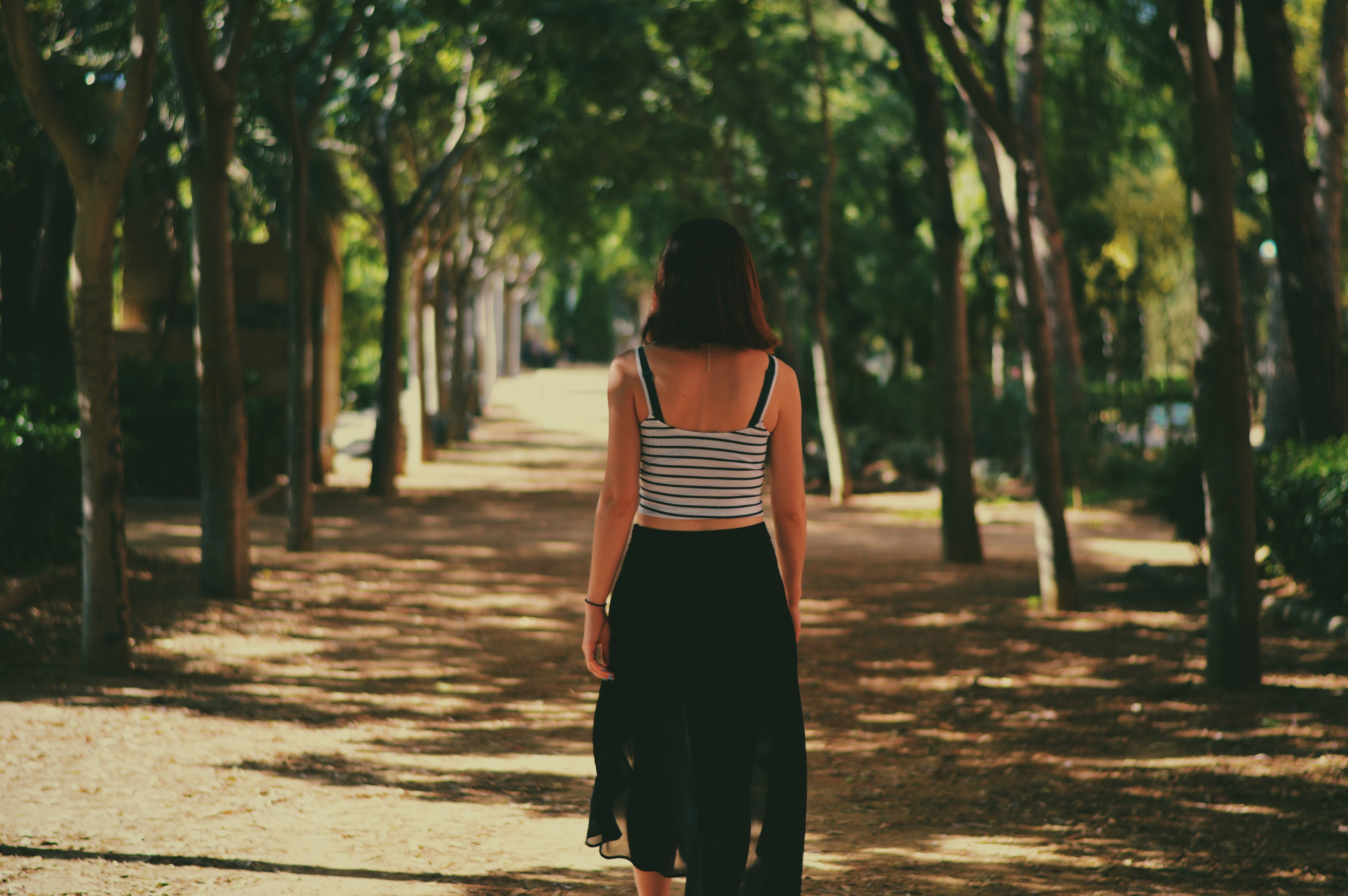 a woman in a striped tank top and black skirt walks away down a tree lined path, woman walking down path 4k, HD Wallpaper