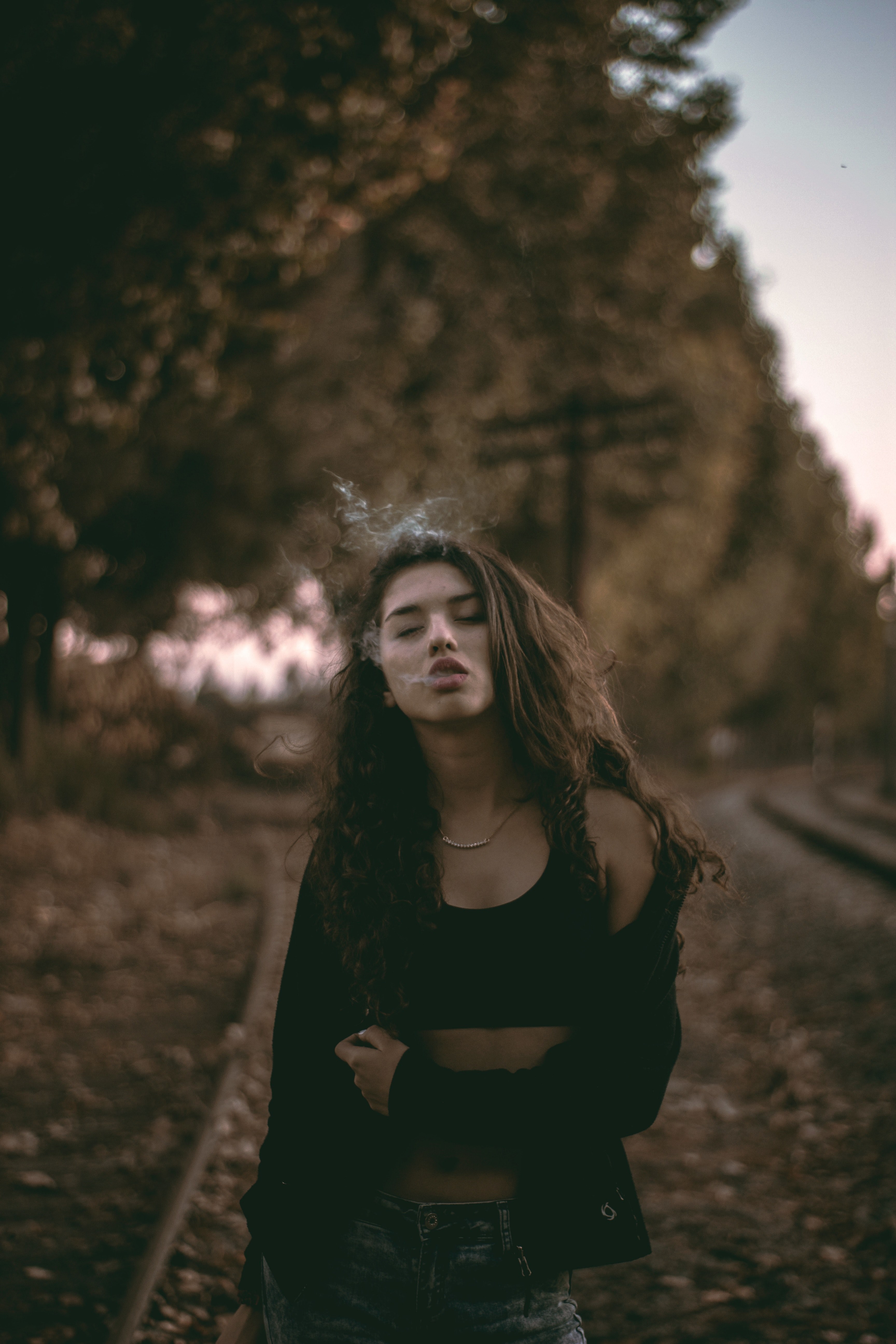 a woman closes her eyes while exhaling smoke outdoors aside train tracks, fran 4k, Phone HD Wallpaper