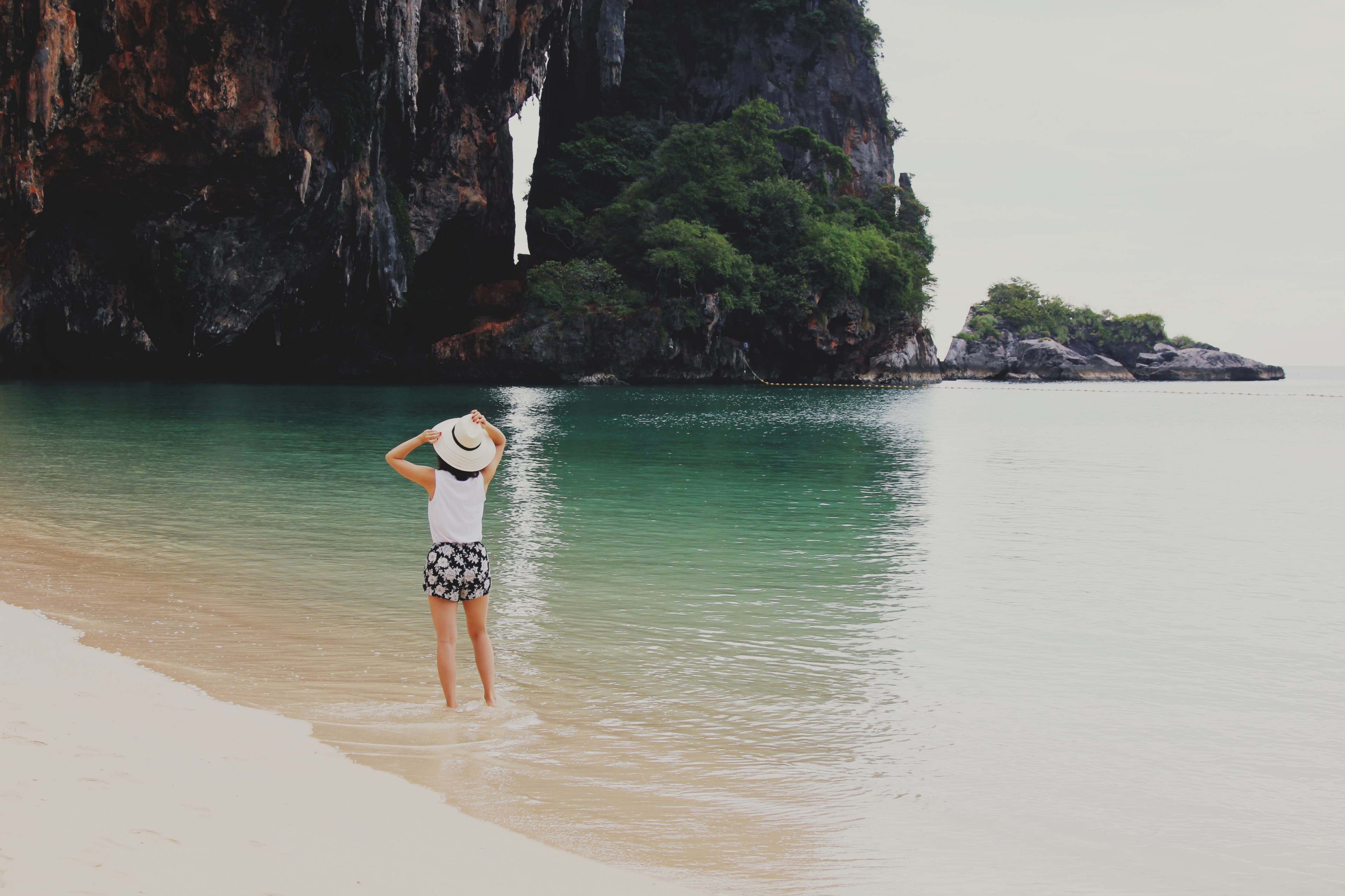 a woman in a hat standing in shallow sea water at the sea shore and looking at the nearby cliff, in the clear water by the cliff 4k, HD Wallpaper
