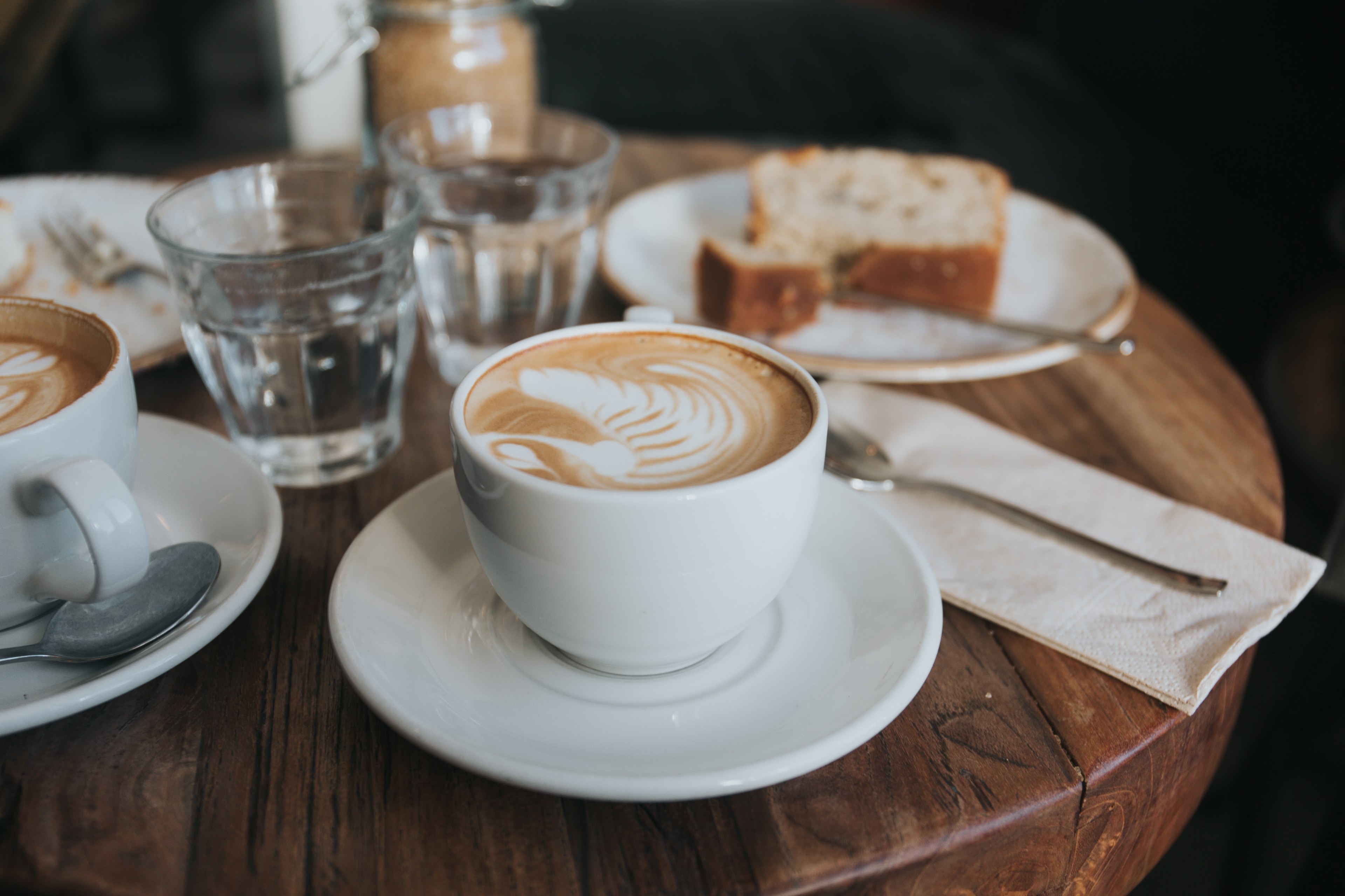 two cappuccinos in white mugs on white saucers with a piece of cake and two water glasses in the background on a wooden table, cappuccinos and cake 4k, HD Wallpaper