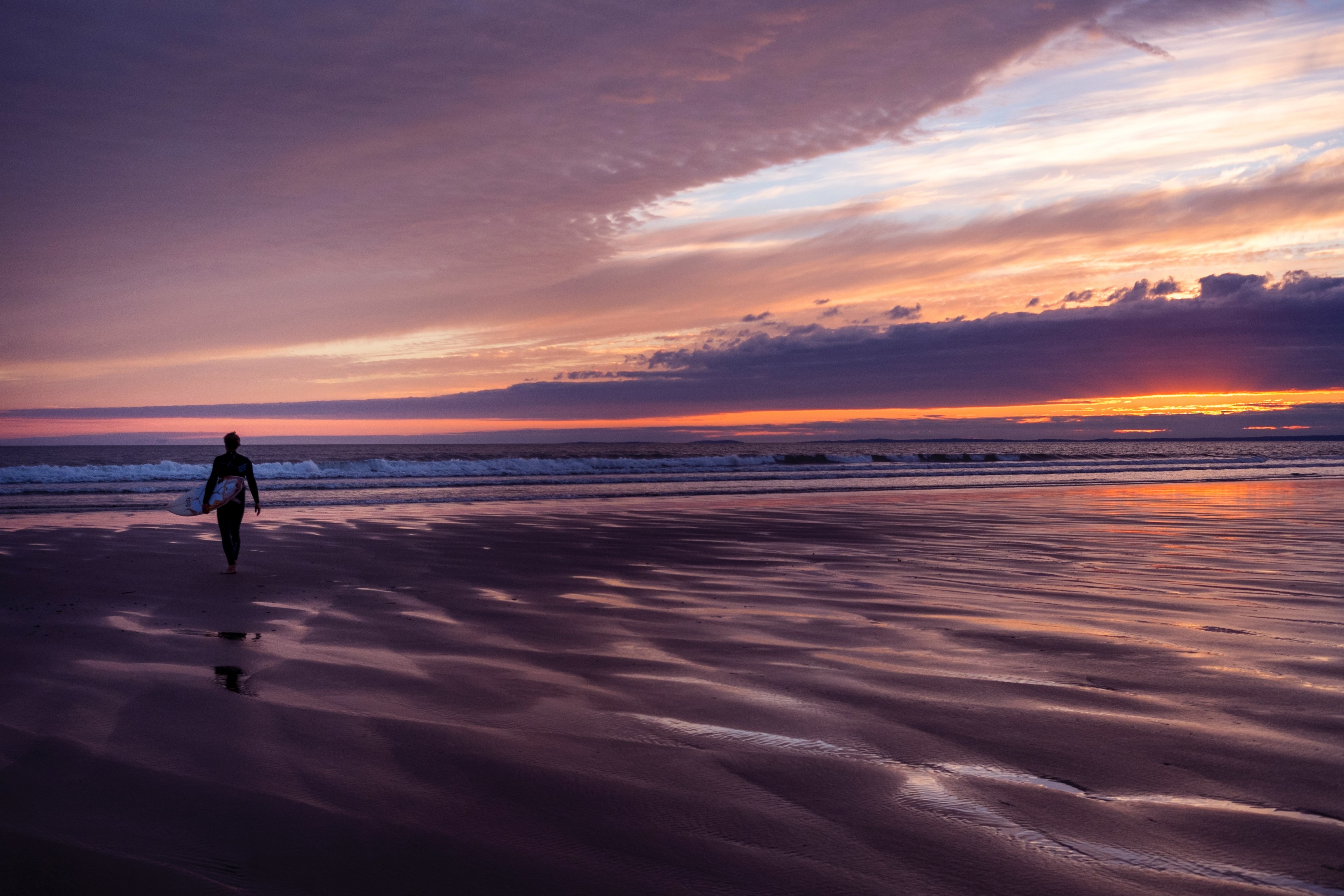 a surfer in a wetsuit walking across a beach at sunset towards the sea with the sun behind a cloud causing a purple sky, beach sunset heavy clouds 4k, HD Wallpaper