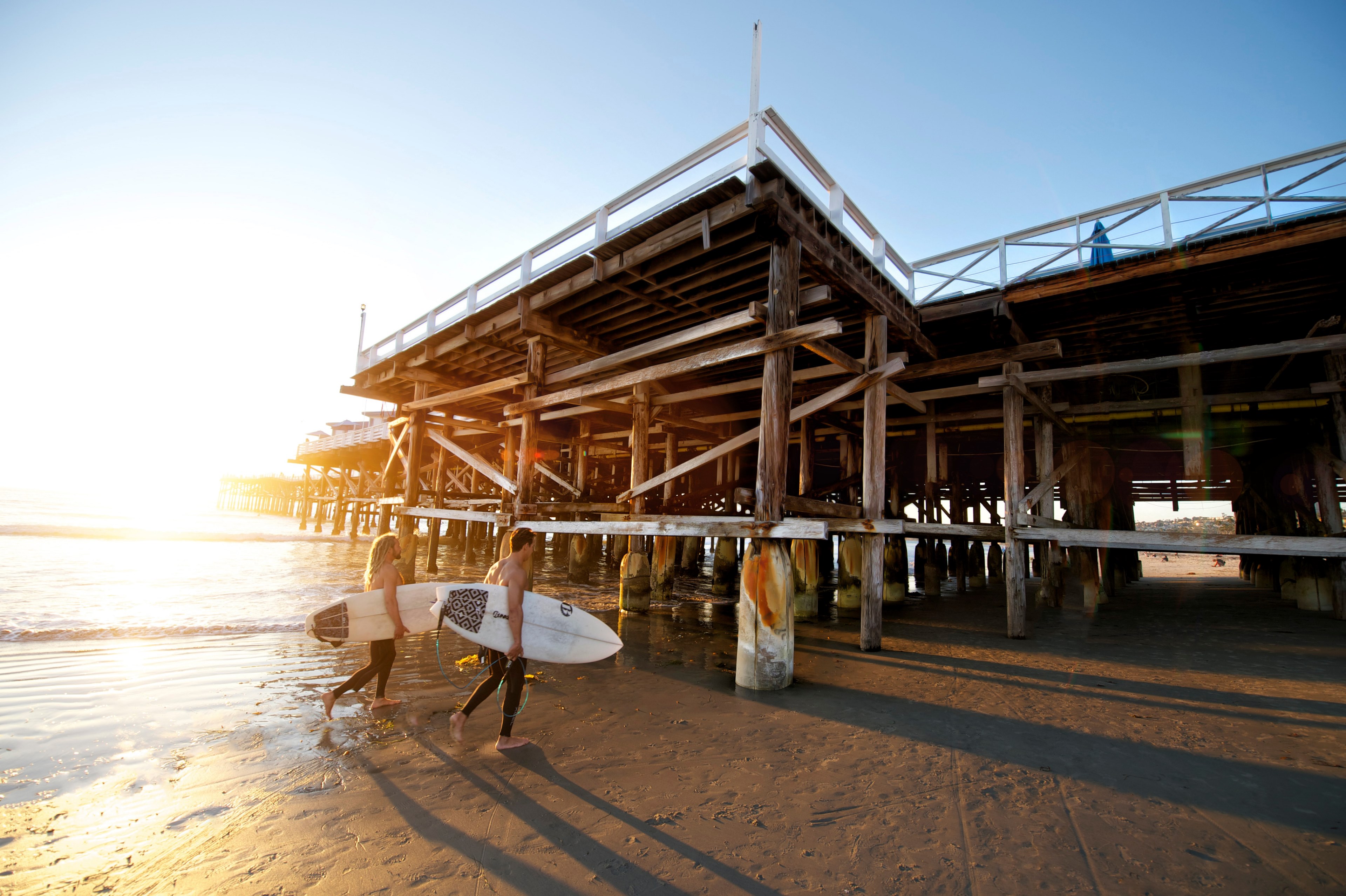 two surfers walking by the wooden pier pillars at sunset, surfers by the pier pillars 4k, HD Wallpaper