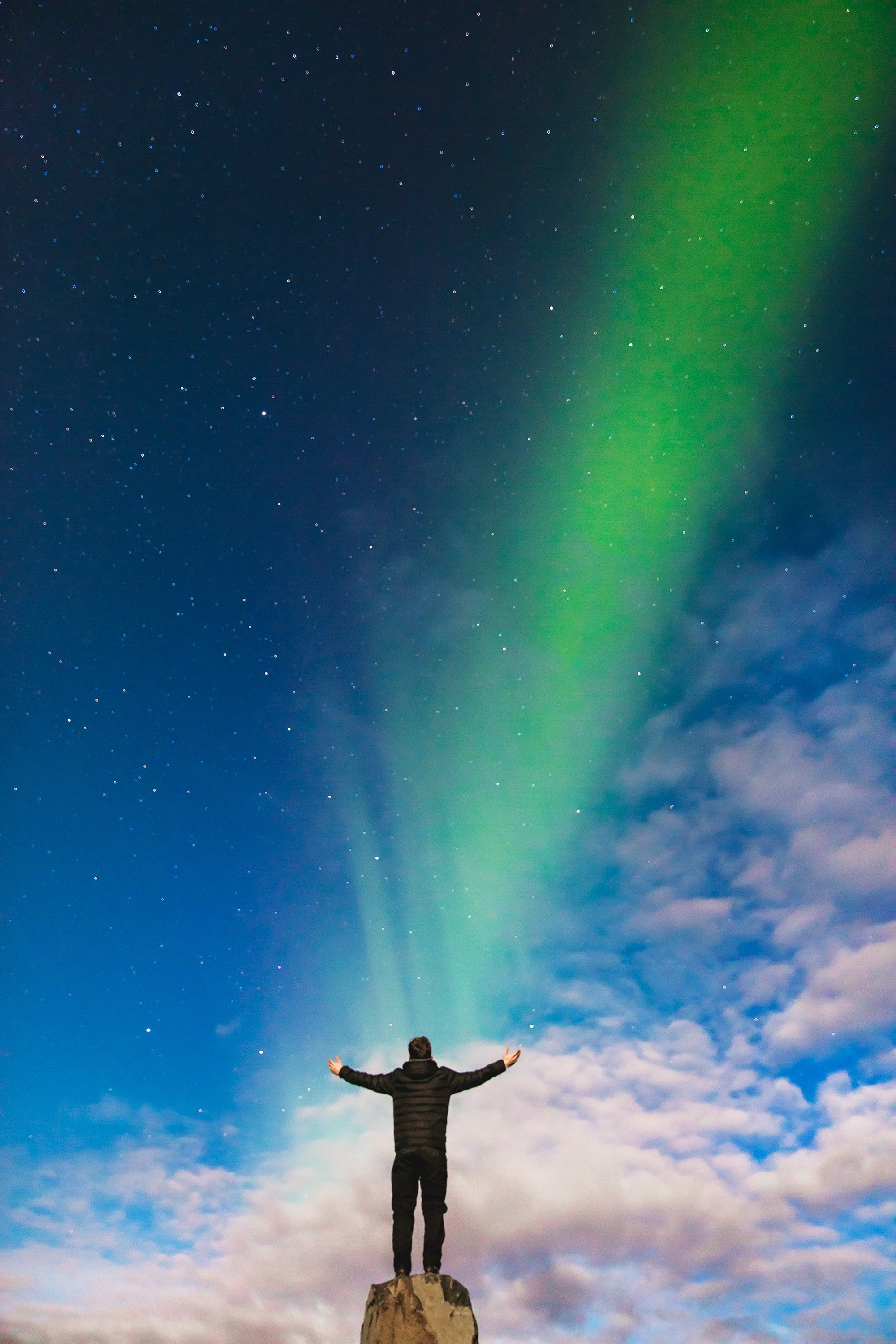 a man raising his arms on top of a rock formation while staring out at a northern lights beam in iceland as it appears to shine onto him, lost in space 4k, Phone HD Wallpaper
