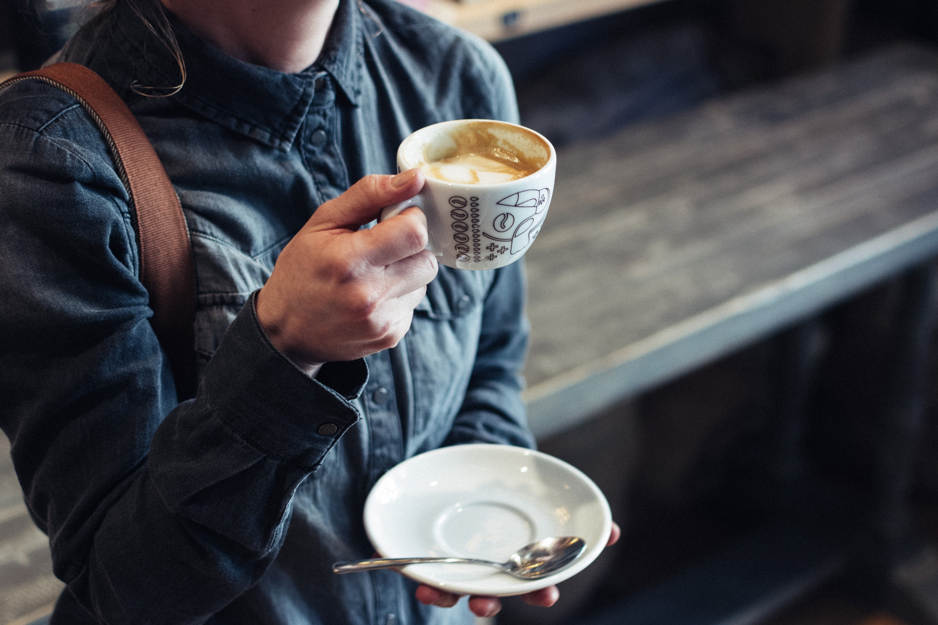 a woman in buttoned up denim shirt with a handbag lifting a mug of cappuccino in one hand and holding a saucer and teaspoon in another, _woman holding her cup of coffee with milk 4k, HD Wallpaper
