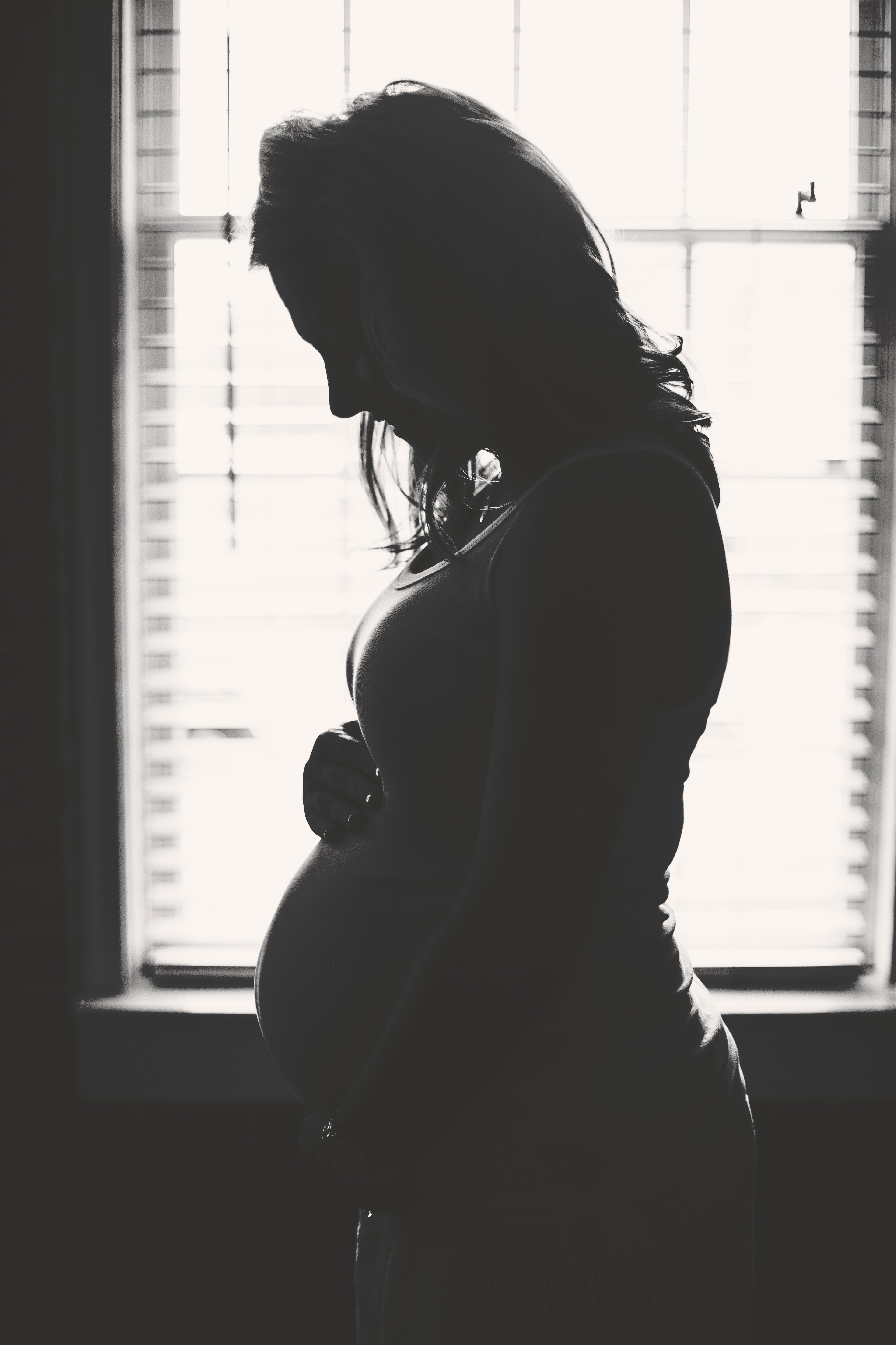 a black and white silhouette of a pregnant woman looking down and holding her belly in front of a window, _a pregnant womans silhouette 4k, Phone HD Wallpaper