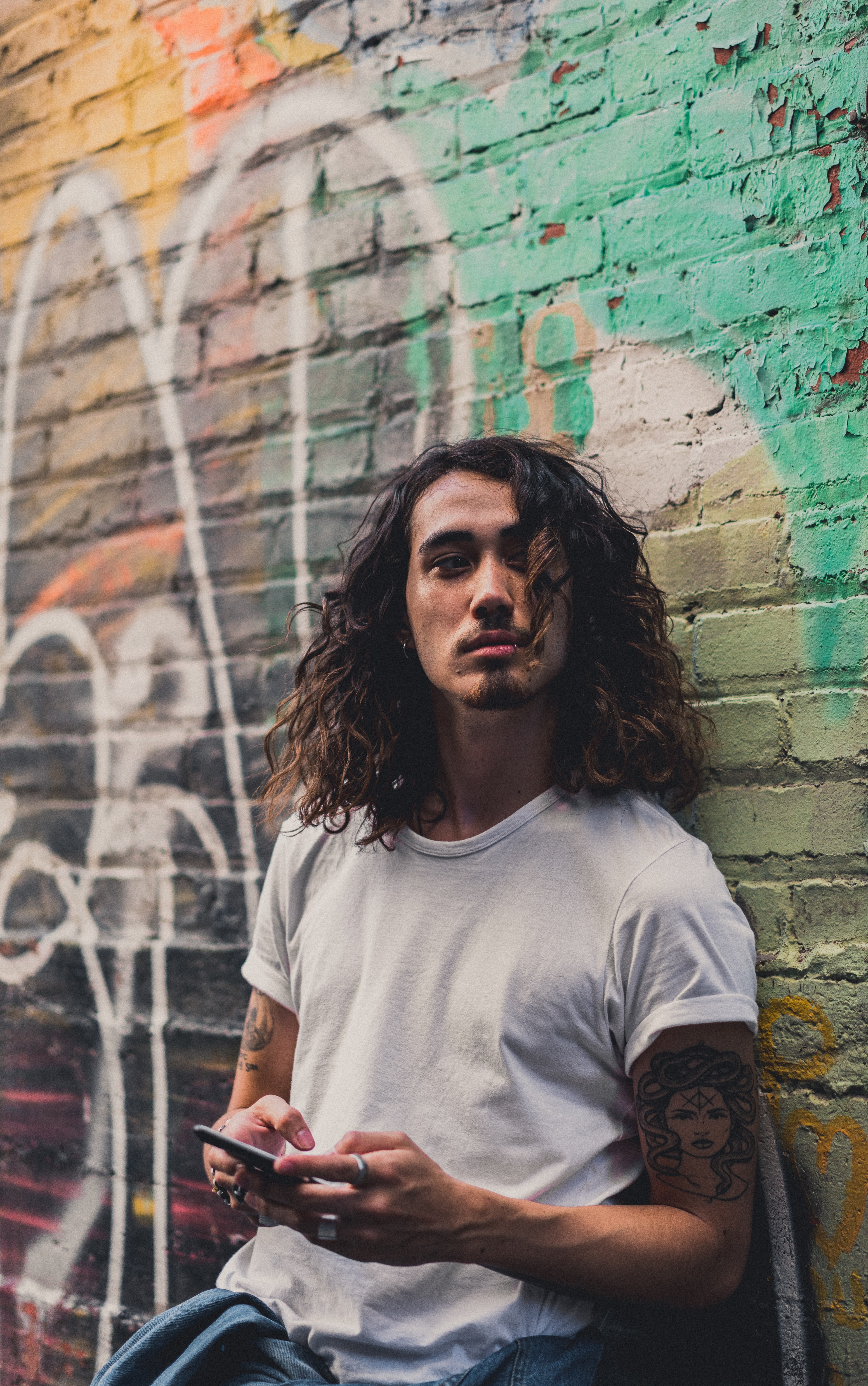 a man with long curly hair leans against a graffitied wall in montreal using an iphone, whos there 4k, Phone HD Wallpaper