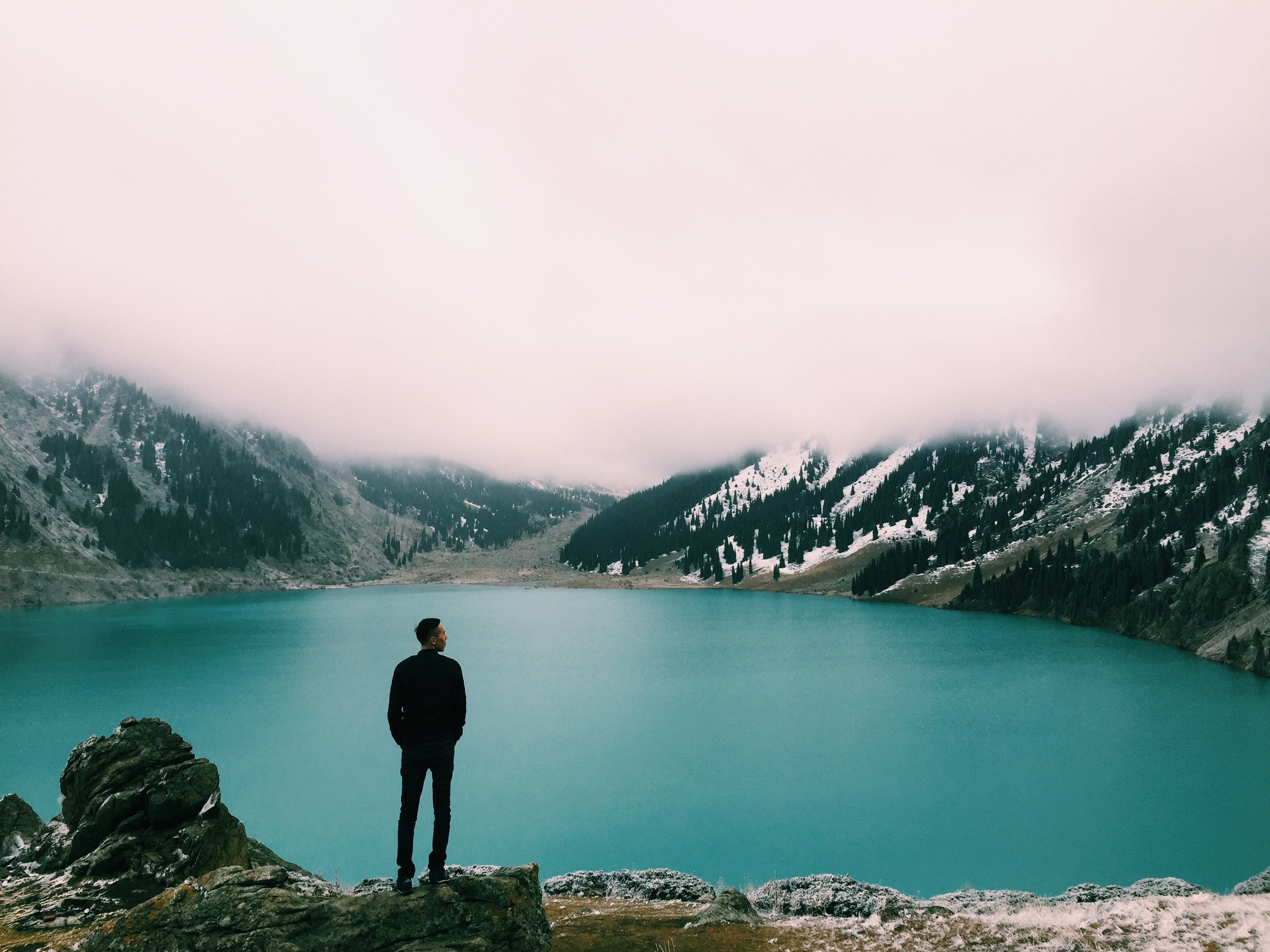 man on a rocky ledge looking at misty mountains and a lake in winter at almaty, man looking at lake almaty 4k, HD Wallpaper