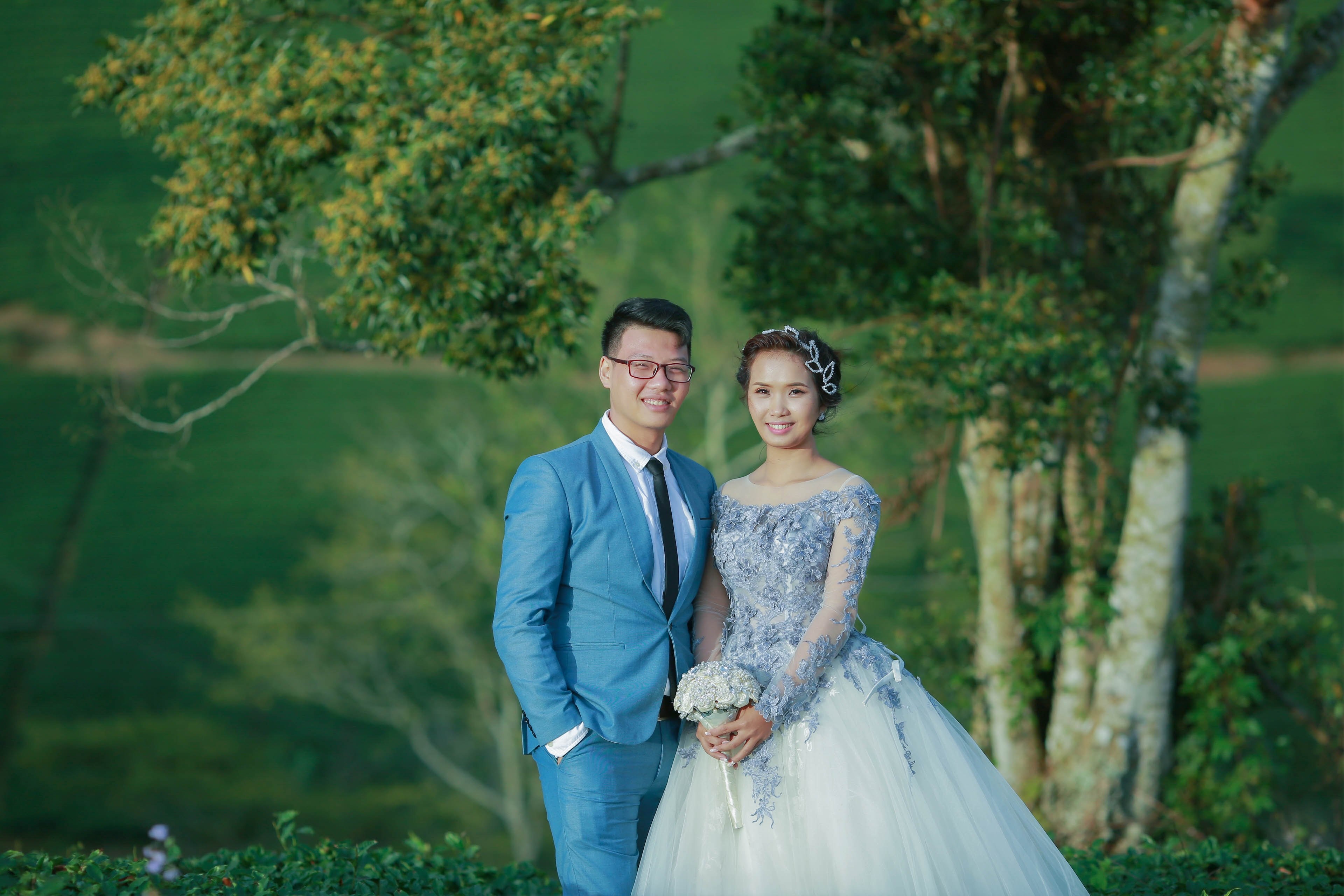 a bride and groom smile at the camera in front of green trees and bushes in vietnam, vietnam wedding 4k, HD Wallpaper