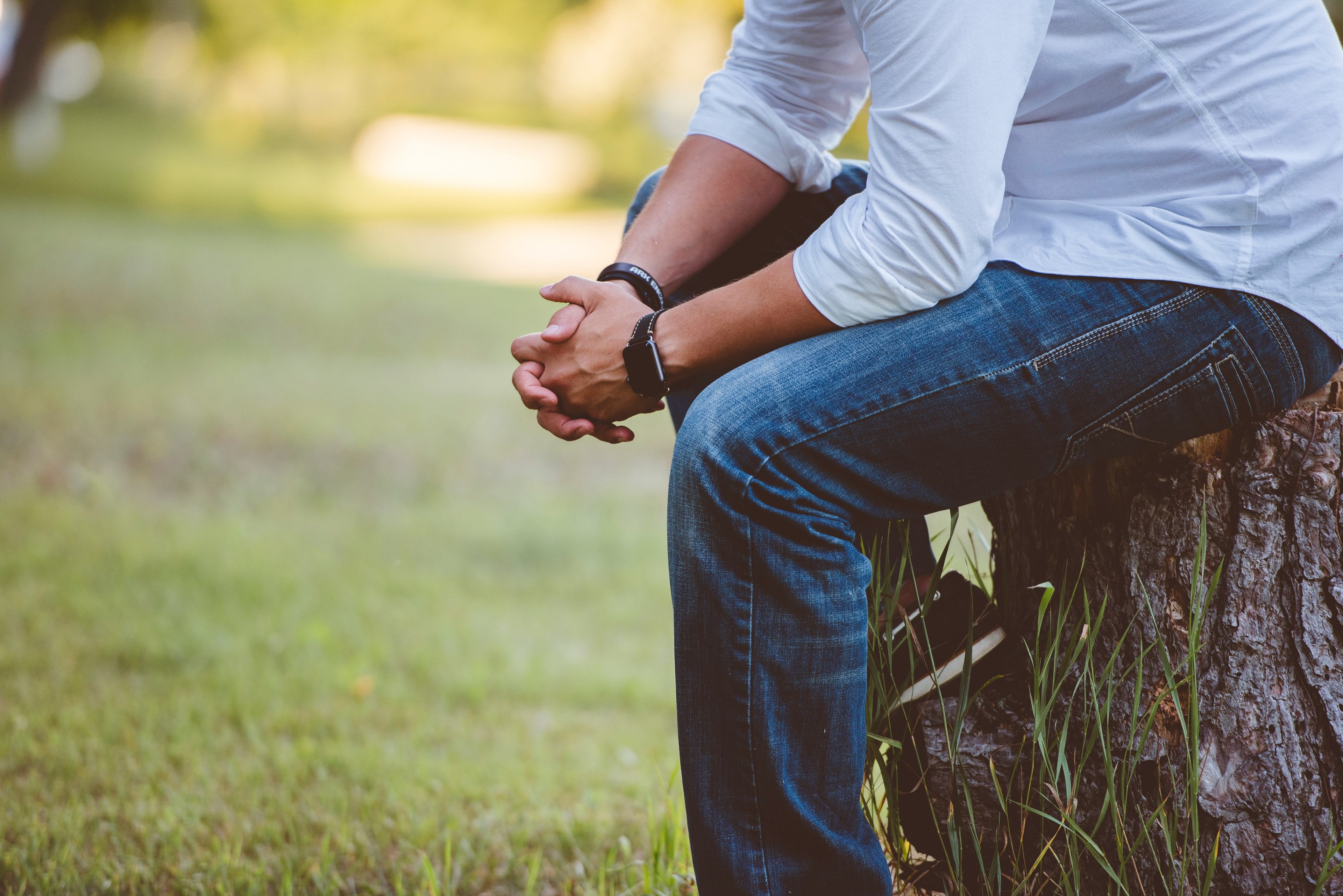 man sitting on stump wearing blue jeans white shirt and a watch with green grass in the background, alone with ones thoughts 4k, HD Wallpaper