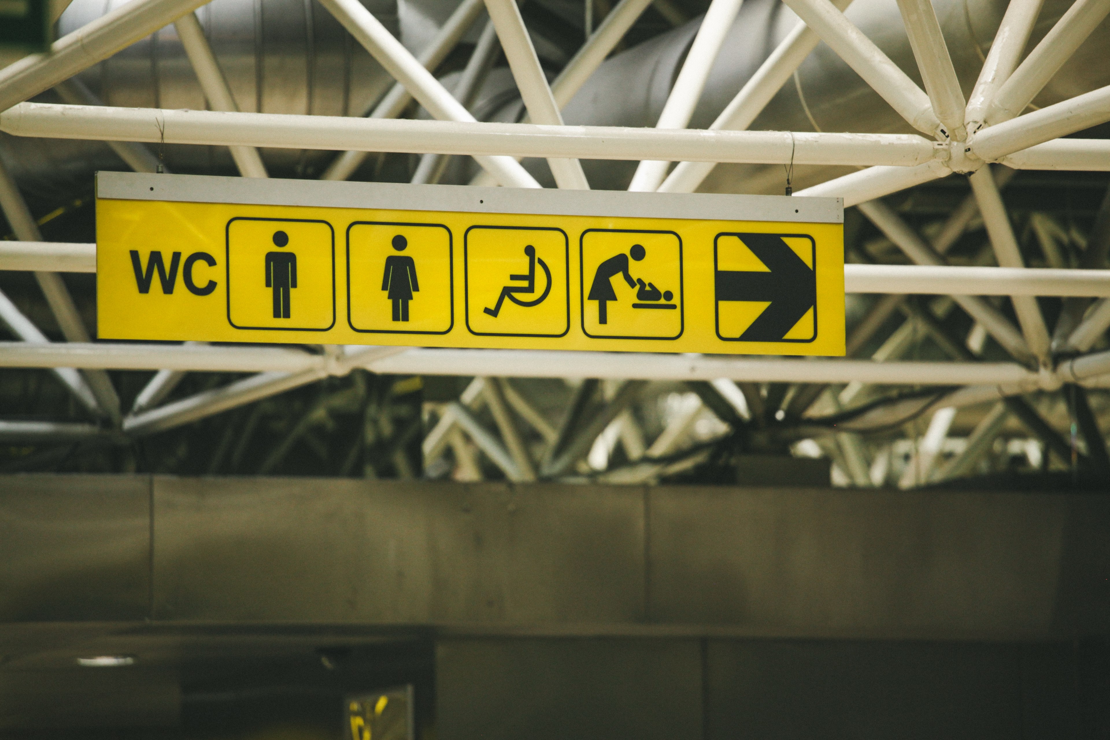 a yellow sign hanging from white scaffolding with the symbols for a male female and handicapped bathroom and baby changing rooms next to an arrow pointing right, yellow airport sign 4k, HD Wallpaper