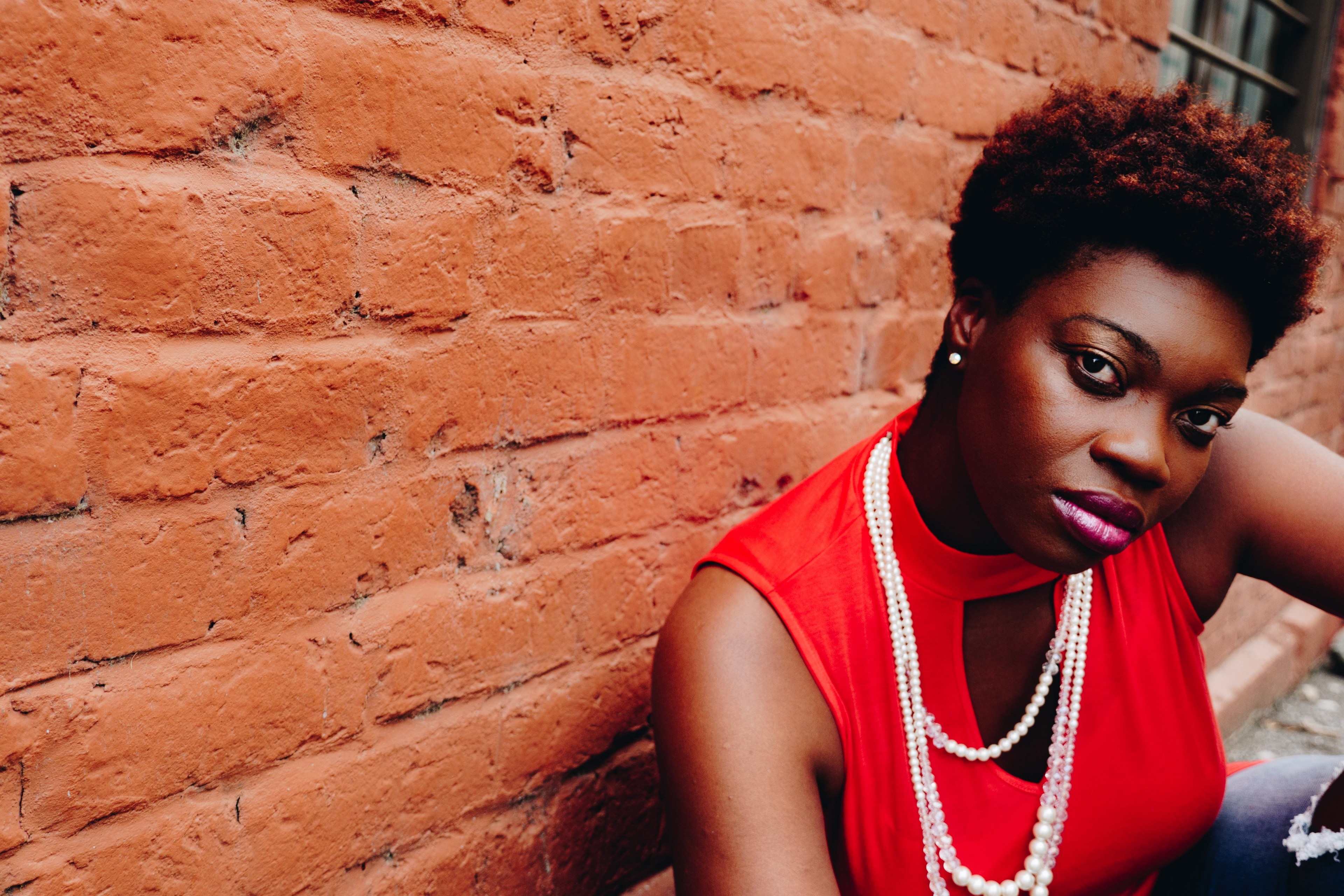 a woman in a long pearl necklace and red shirt sits by a red brick wall, woman in long pearl necklace 4k, HD Wallpaper