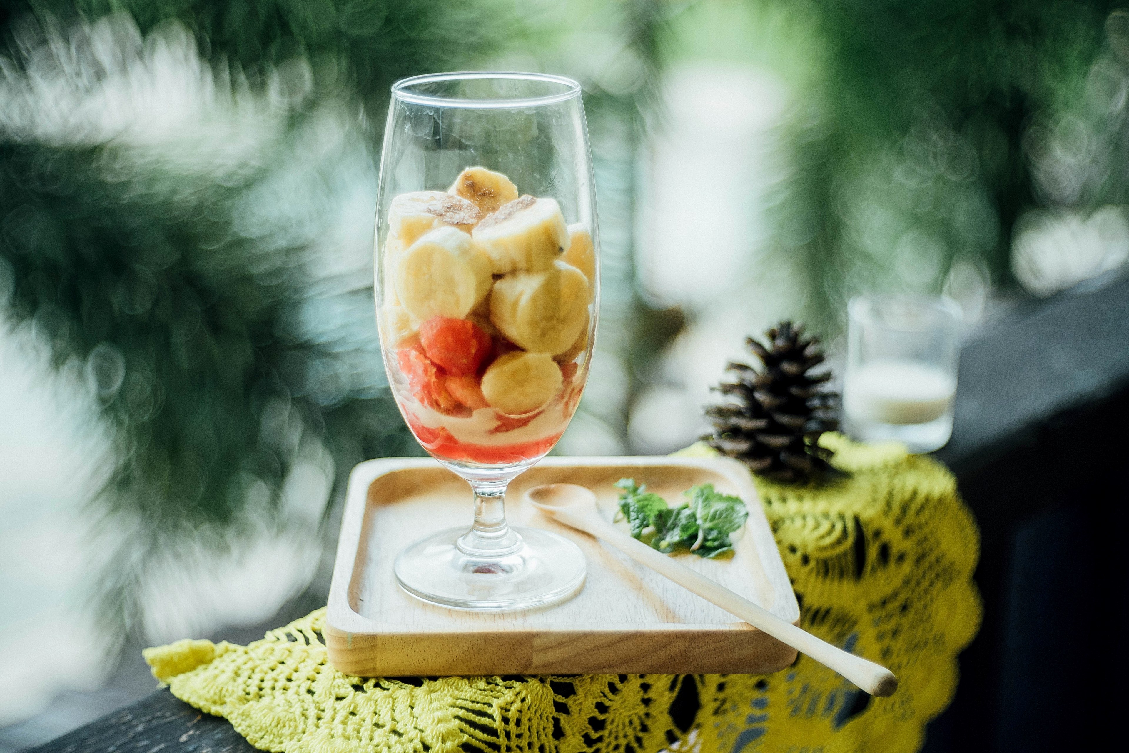 a glass of chopped bananas and strawberries beside a long wooden spoon and mint on a wood coaster on top of a yellow doily next to a pinecone and a glass of milk, my healthy food 4k, HD Wallpaper