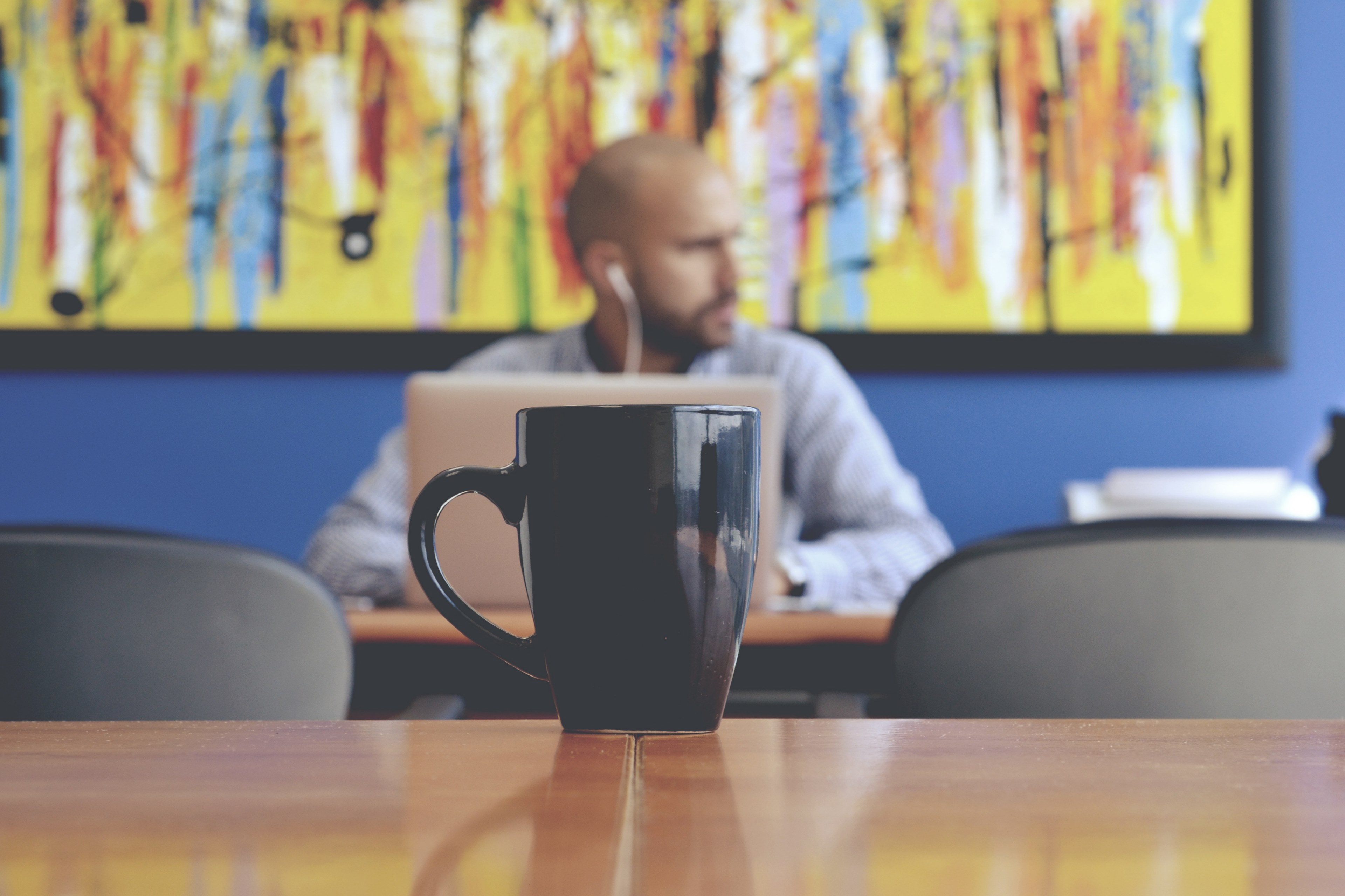a fuzzy shot of a mug on a table and a man sitting at a laptop at the back of the room, working man behind a mug 4k, HD Wallpaper