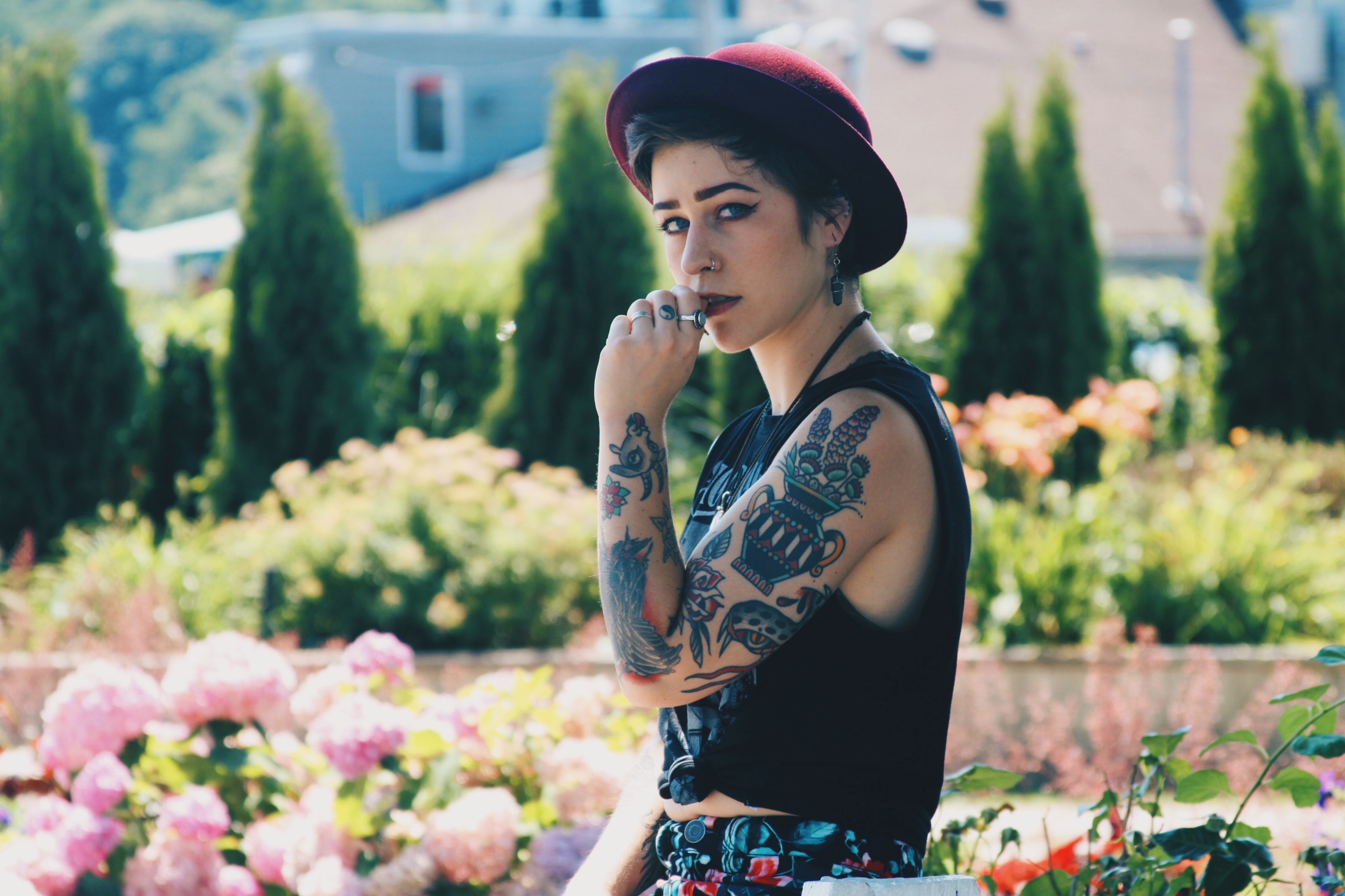 a tattooed woman in a red hat stands before a lush flower garden, woman in front of garden 4k, HD Wallpaper