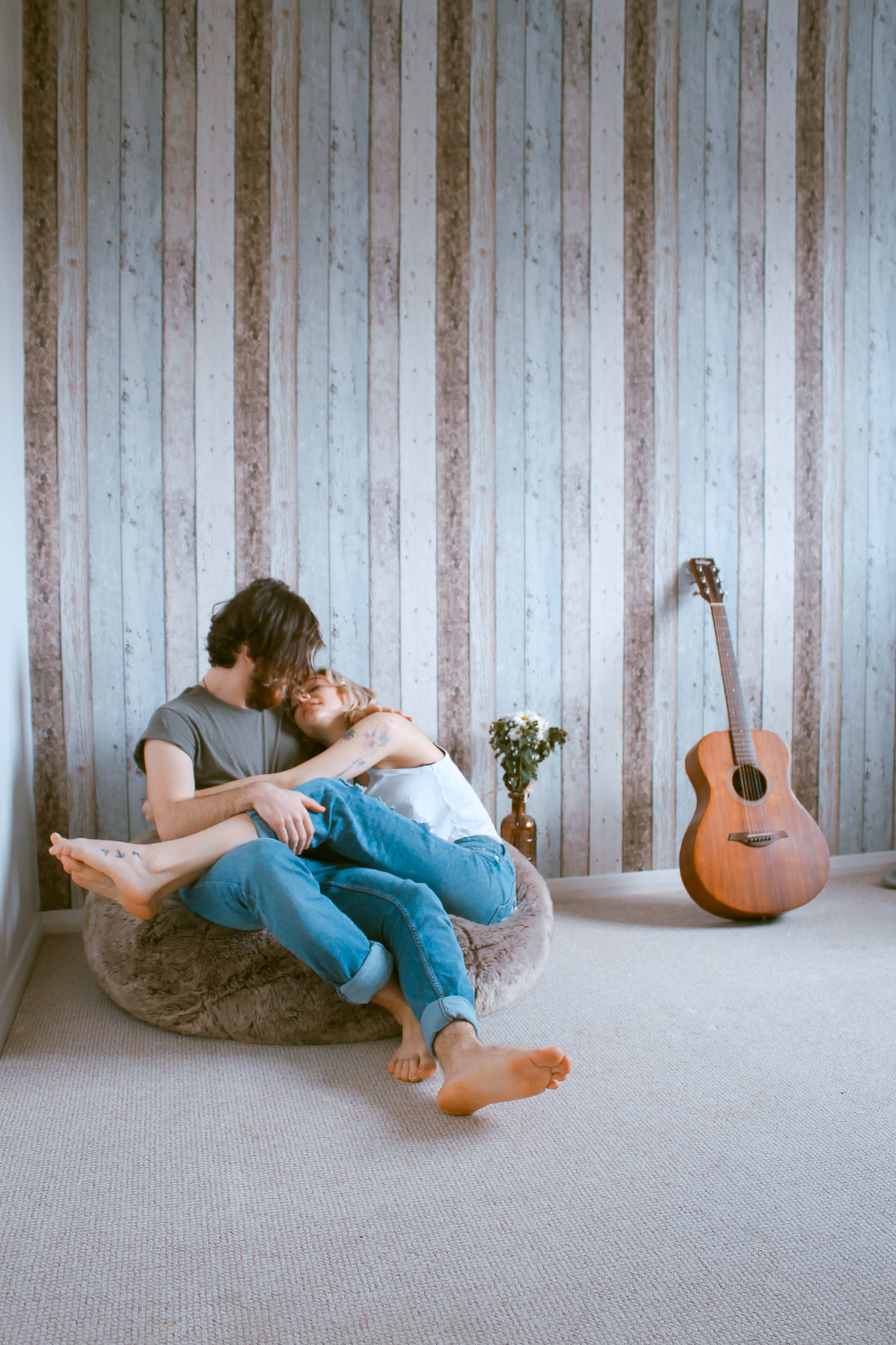 a barefooted man and woman cuddle on a beanbag chair against wood paneling near a guitar, still in your arms 4k, Phone HD Wallpaper