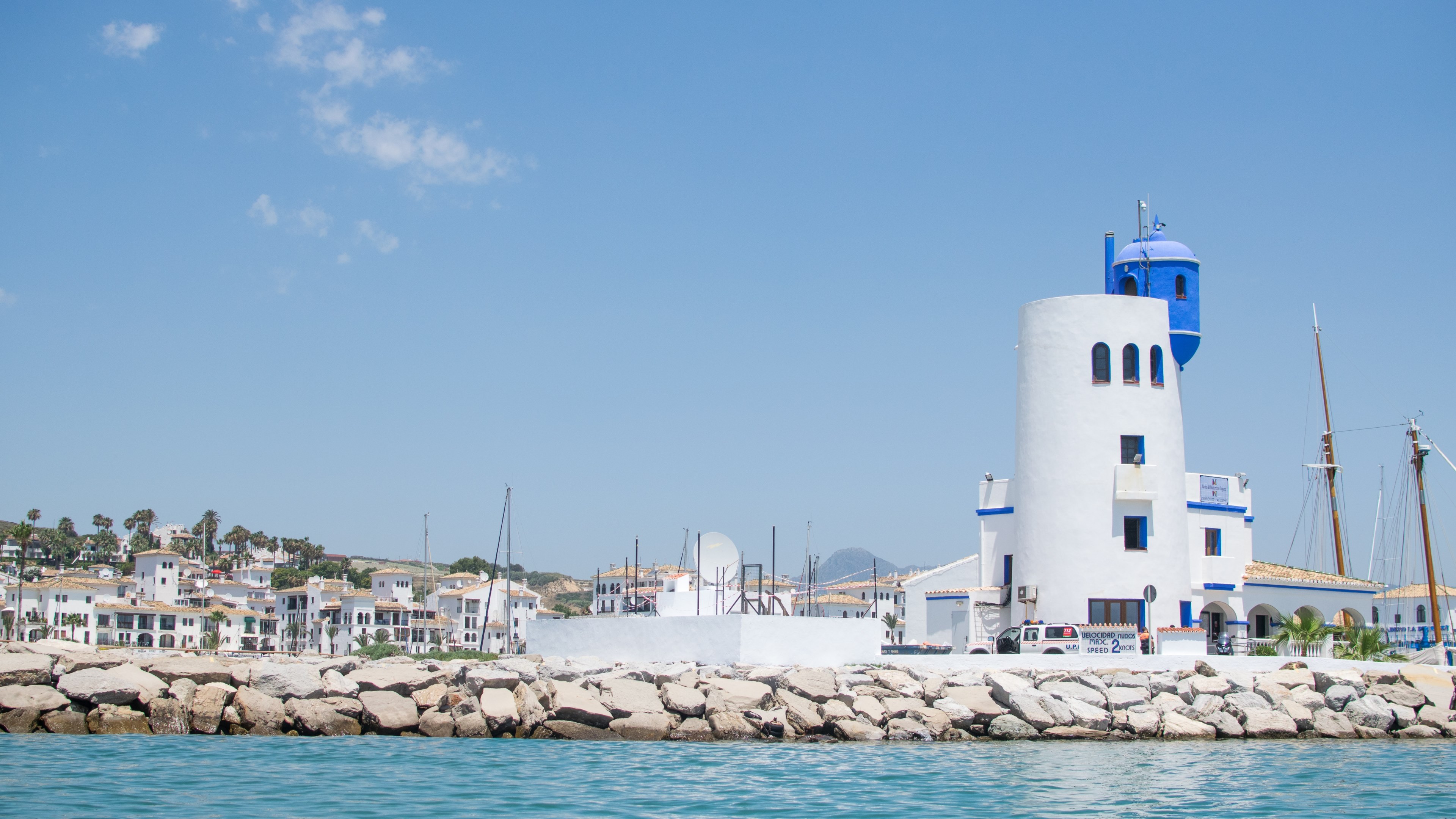 a white and blue lighthouse on the waterfront of a mediterranean town on a sunny day, the lighthouse 4k, HD Wallpaper