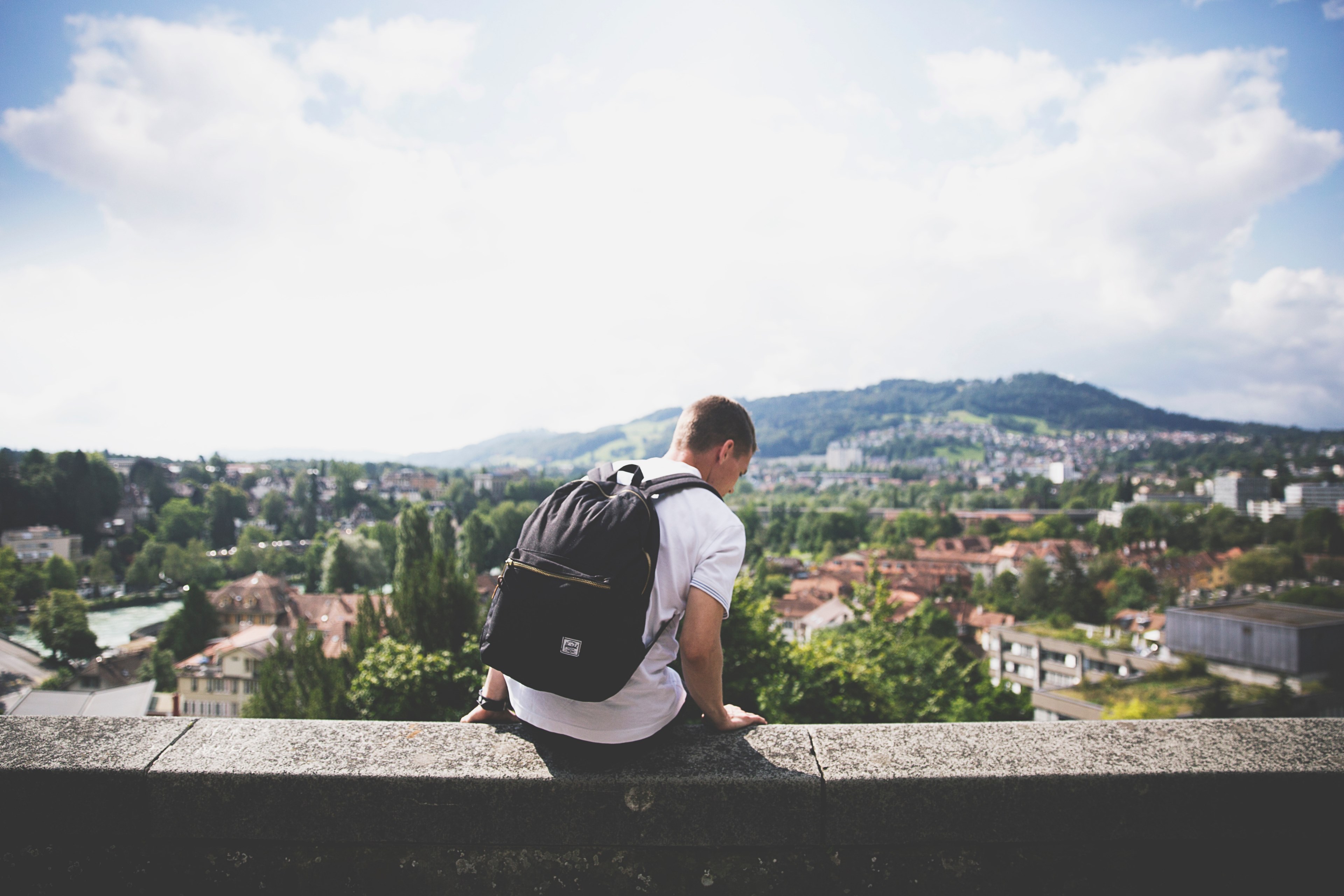 a man with a backpack sitting on a ledge overlooking a picturesque town, vacation in a pleasant town 4k, HD Wallpaper