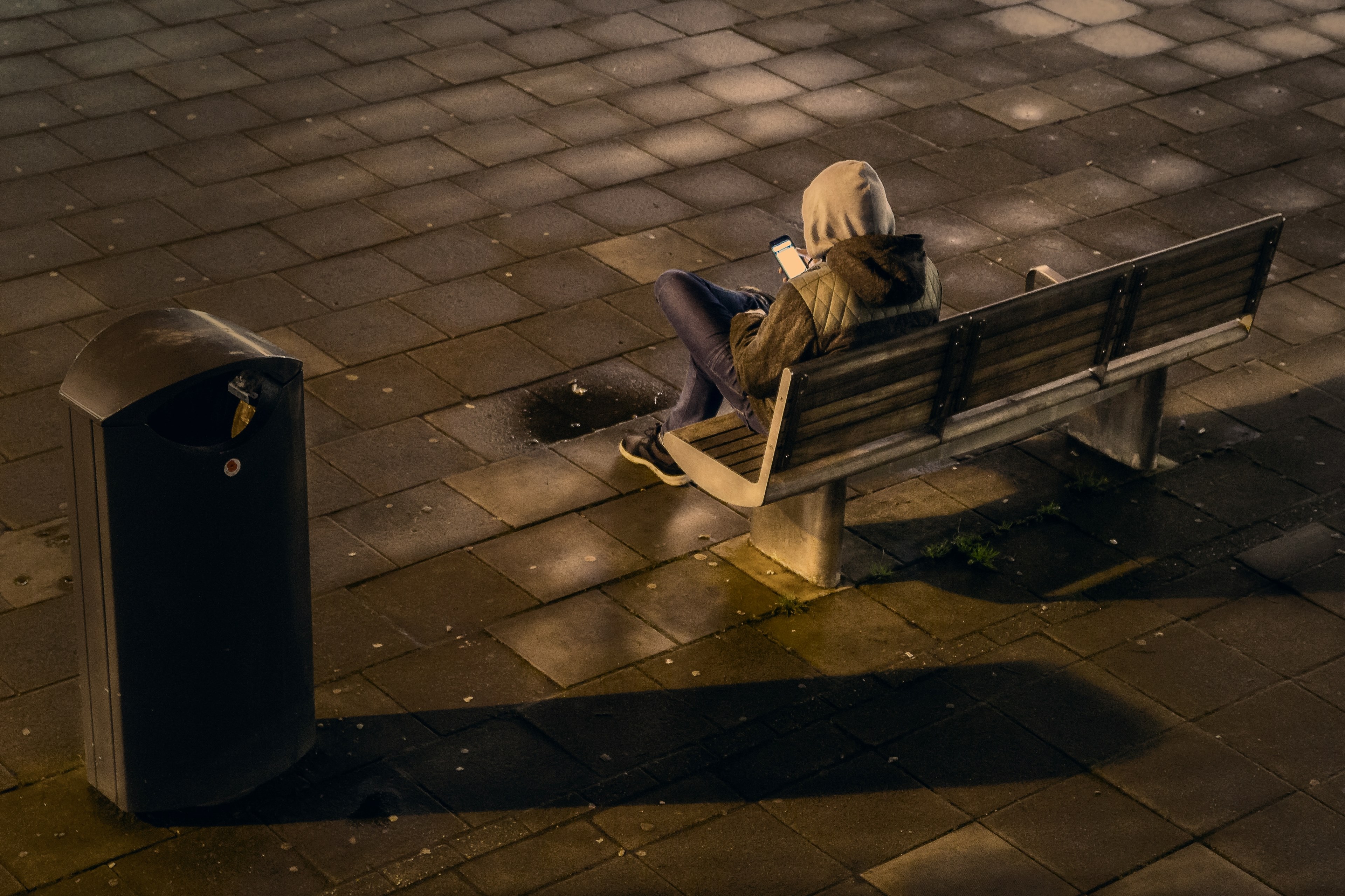 a hooded man on a bench using his smart phone beside a garbage can in a tile floor outside at night, hooded man on bench 4k, HD Wallpaper
