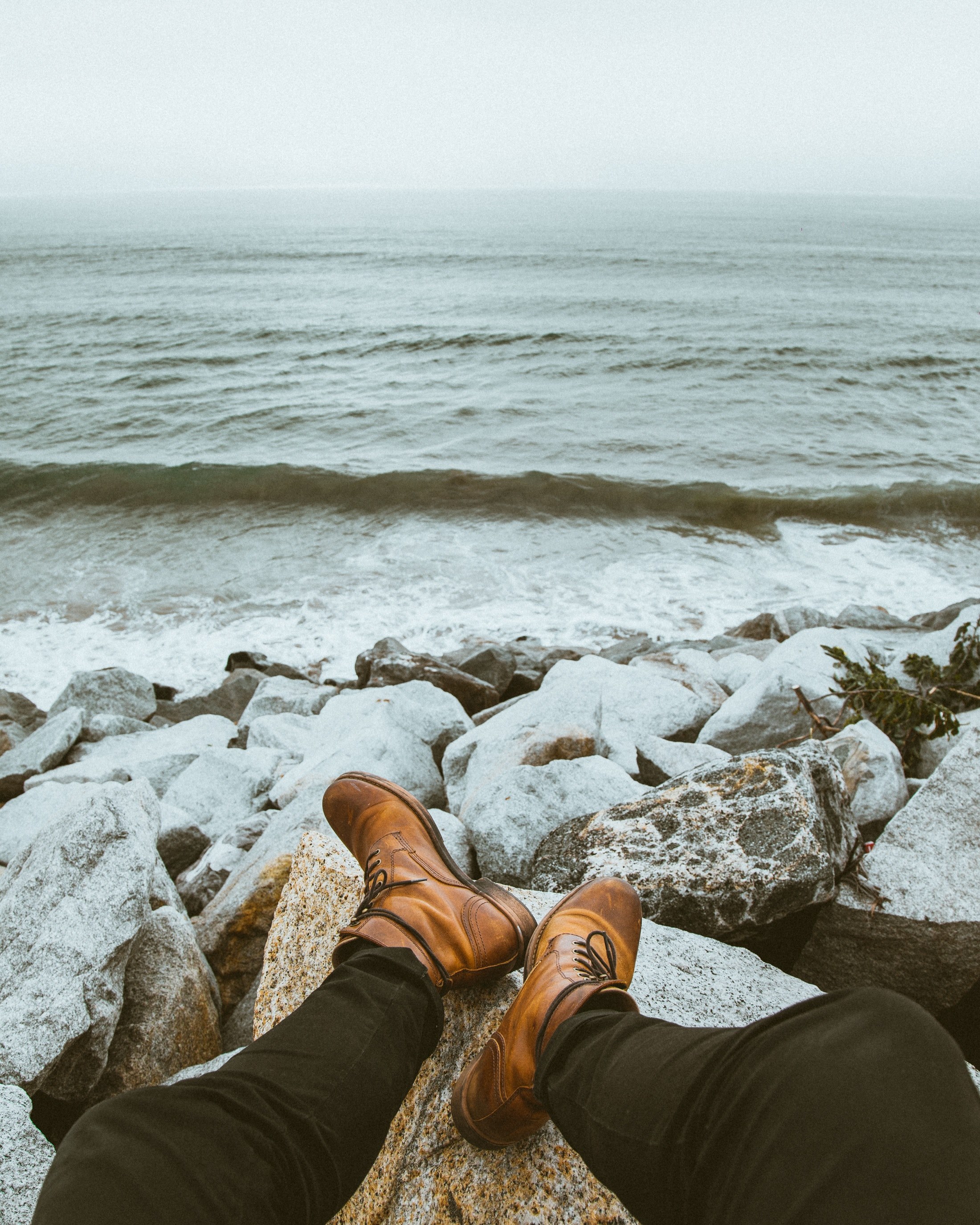 a person wearing black jeans and boots sitting on rocks near the water, waiting for sunset 4k, Phone HD Wallpaper