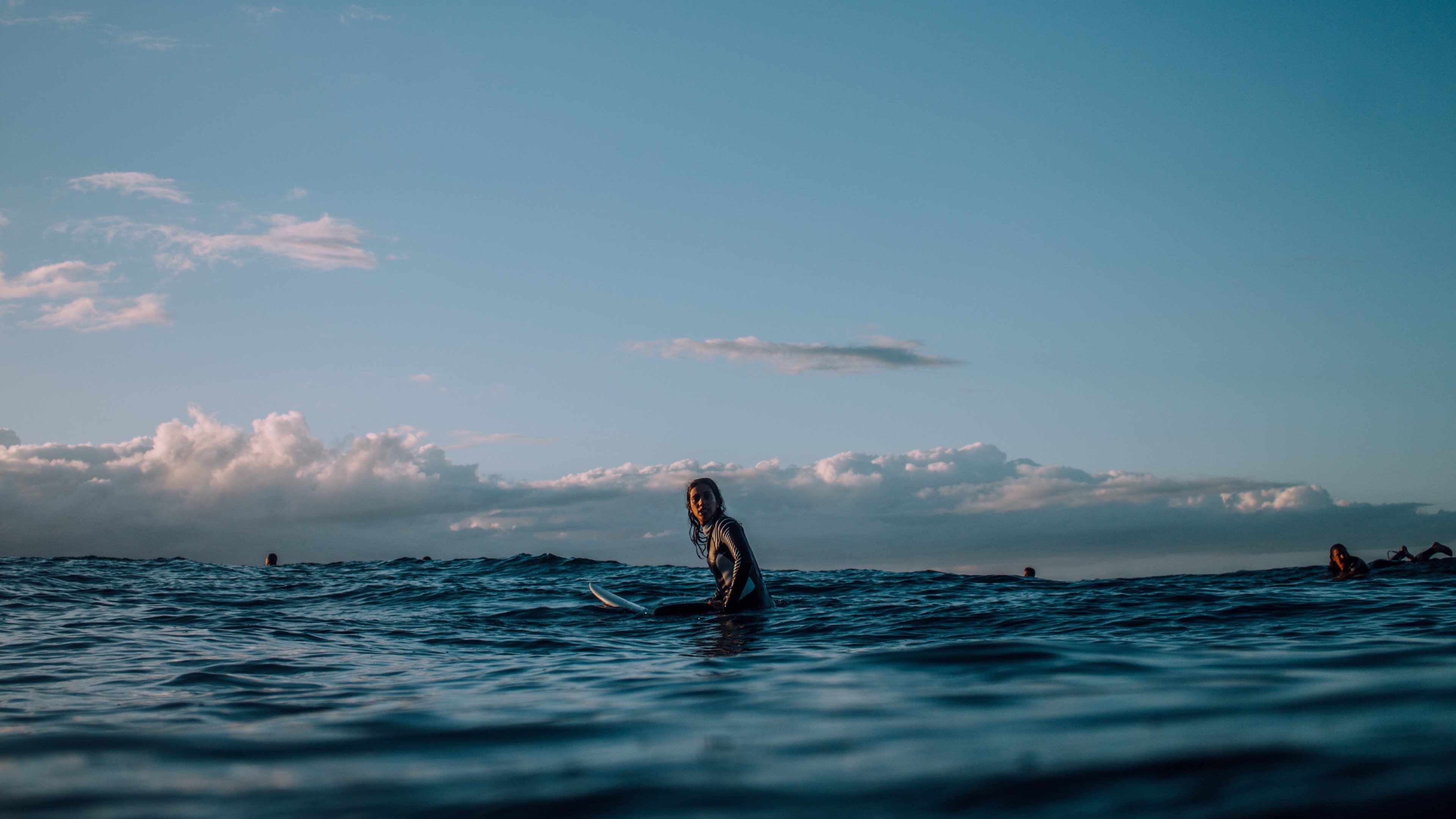 a female surfer sat on her board in the calm water and under a blue cloudy sky at piha with other surfers in the background, should be a corona ad 4k, HD Wallpaper