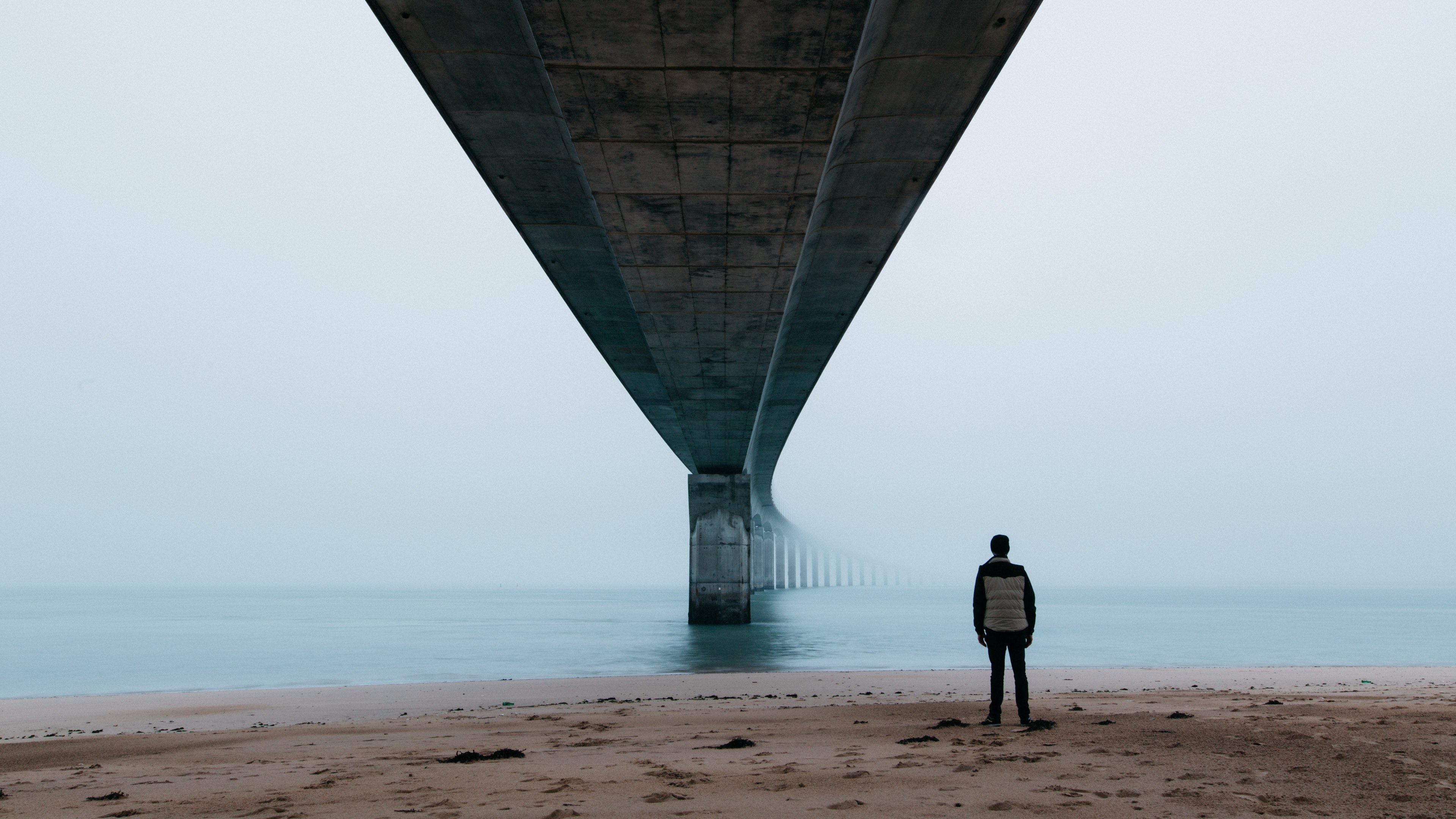 a man standing on the beach under a bridge looking out into an ocean covered in fog and mist, foggy day_ 4k, HD Wallpaper