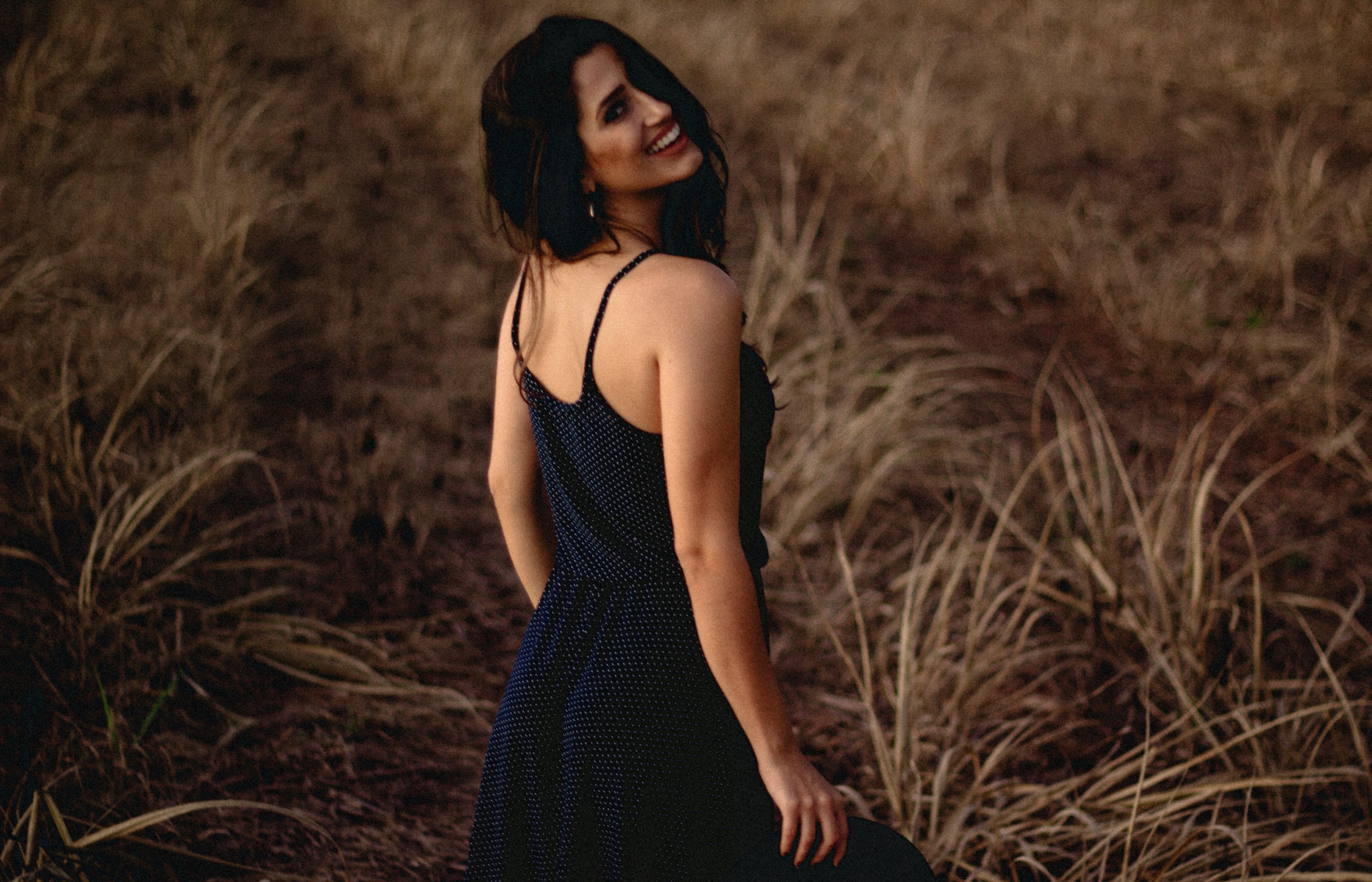 a woman in a black dress smiles over her shoulder in a field of brown grass, woman smiling over shoulder 4k, HD Wallpaper
