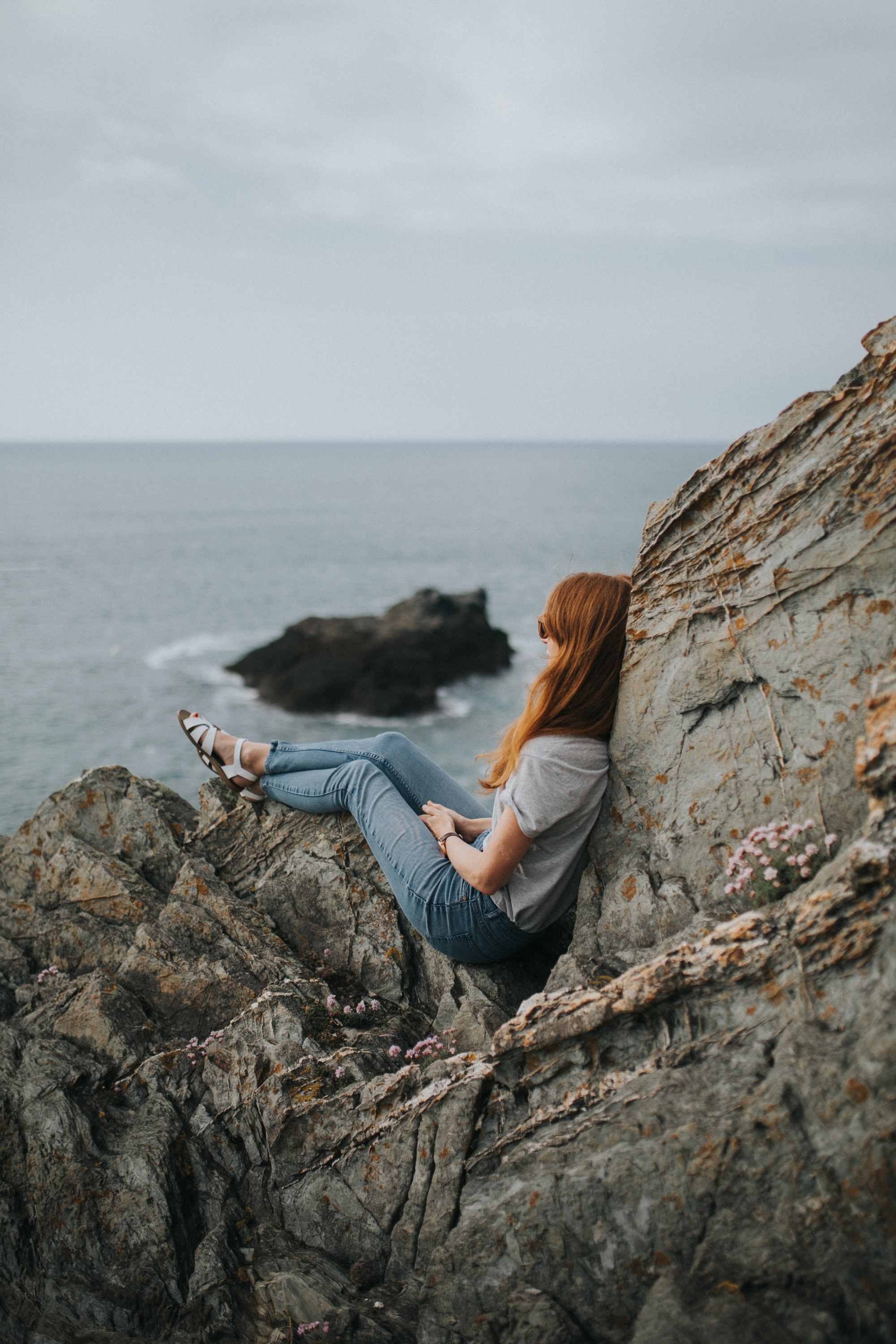 woman in jeans and shirt sitting on coastline rocks at fistral beach, woman sitting on the rocks 4k, Phone HD Wallpaper