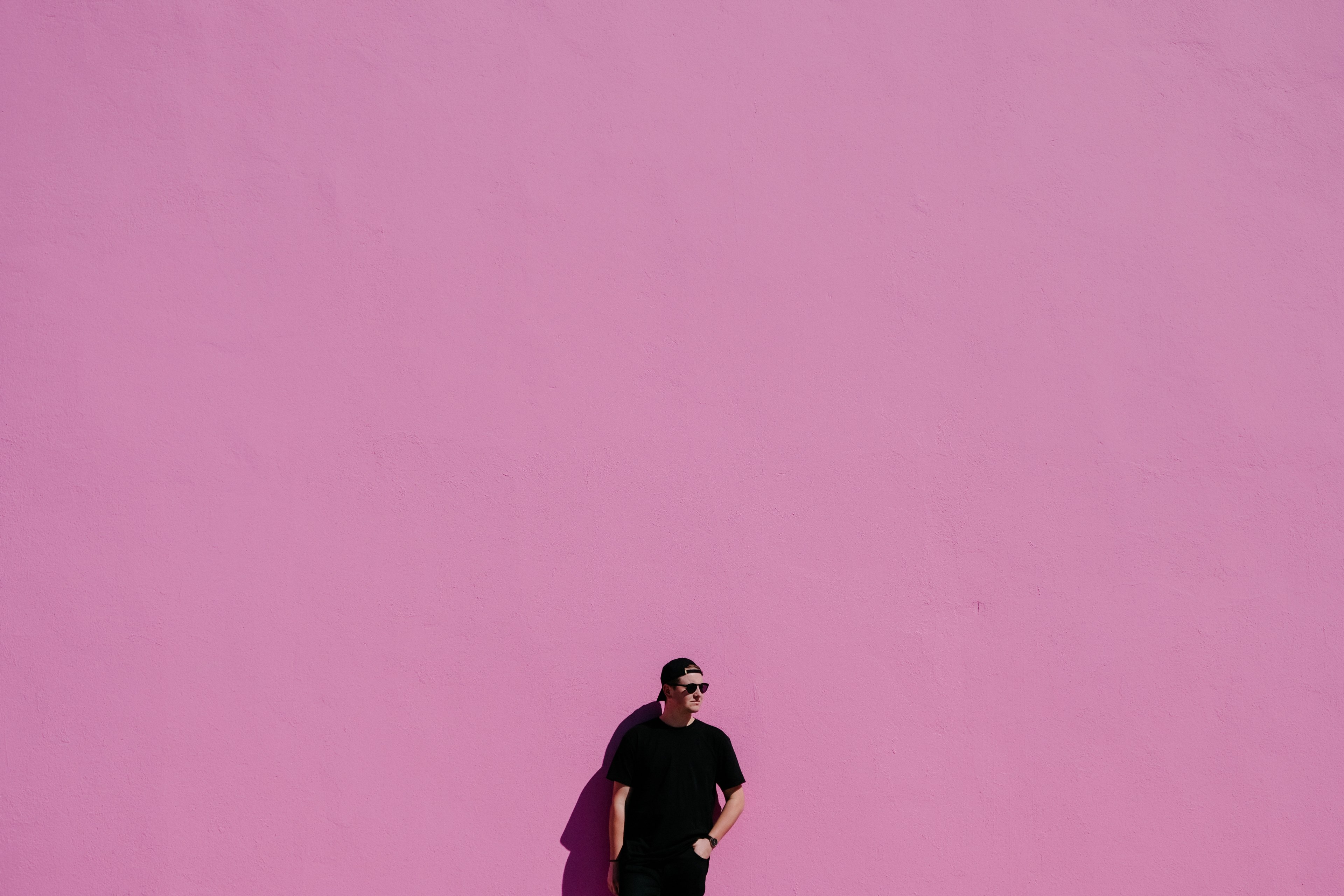 a man in a backwards baseball cap black t shirt and watch stands against a pink wall, man against pink wall 4k, HD Wallpaper