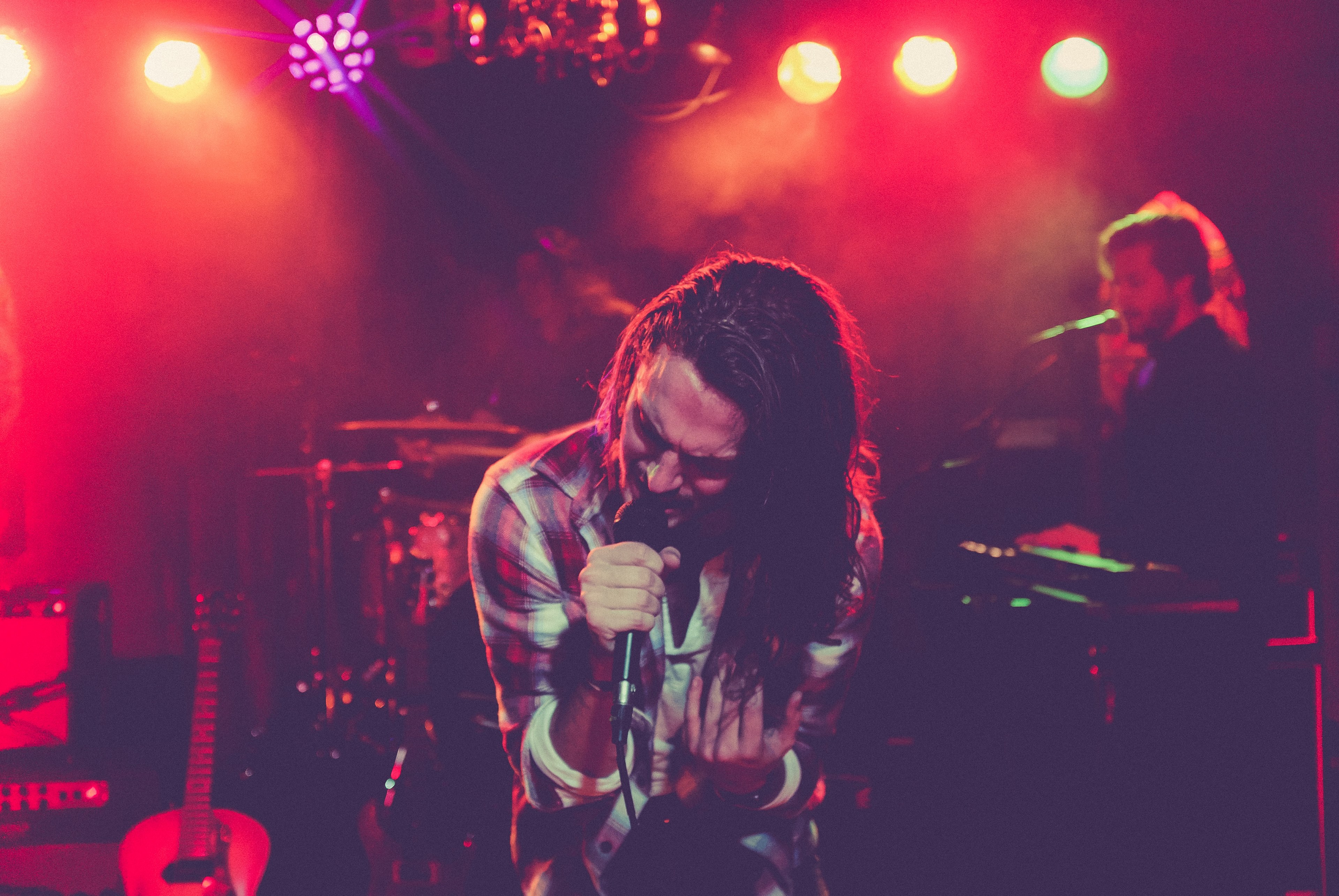 a long haired male vocalist in a checkered shirt sings into the microphone under red floodlights, expressive vocalist on stage 4k, HD Wallpaper