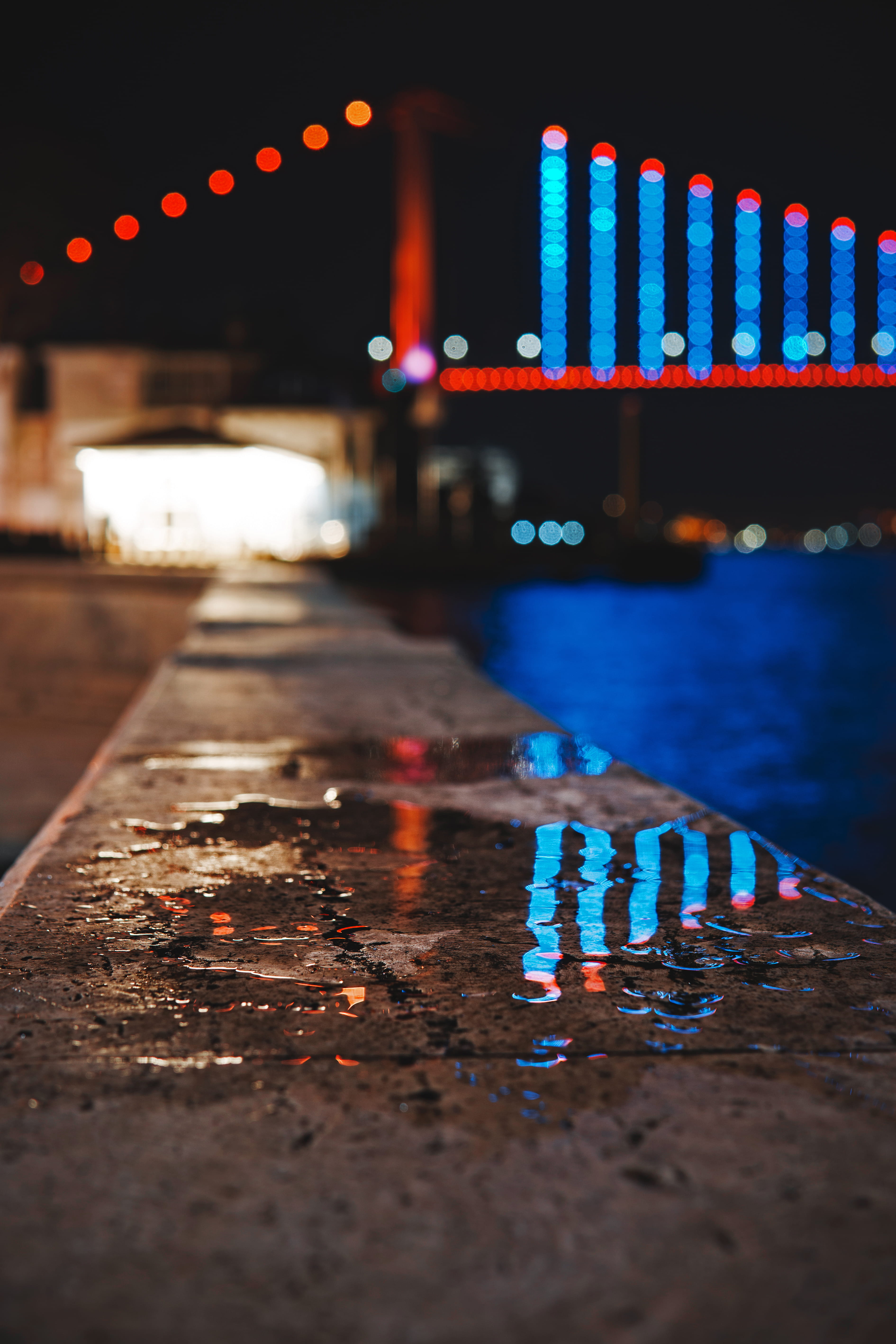 selective focus, surface level, dark, water, outdoors, building exterior, blur, transportation, pavement, lights, bridge, lighting equipment, lighted, evening, Phone HD Wallpaper