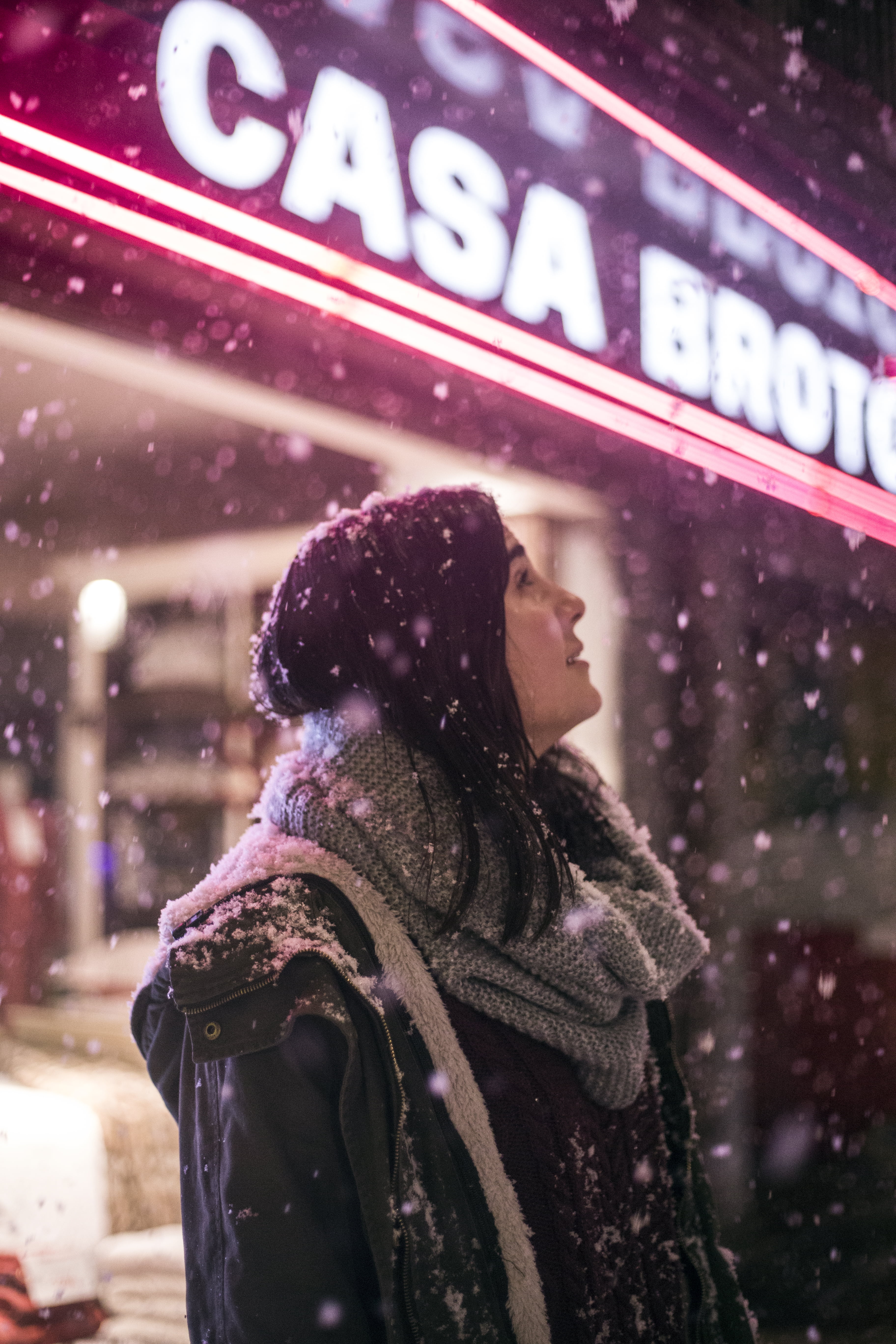 focus on foreground, snow flakes, snow, city, real people, blur, street, sign, winter coat, wear, warm clothing, happy, woman, hood - clothing, close-up, Phone HD Wallpaper