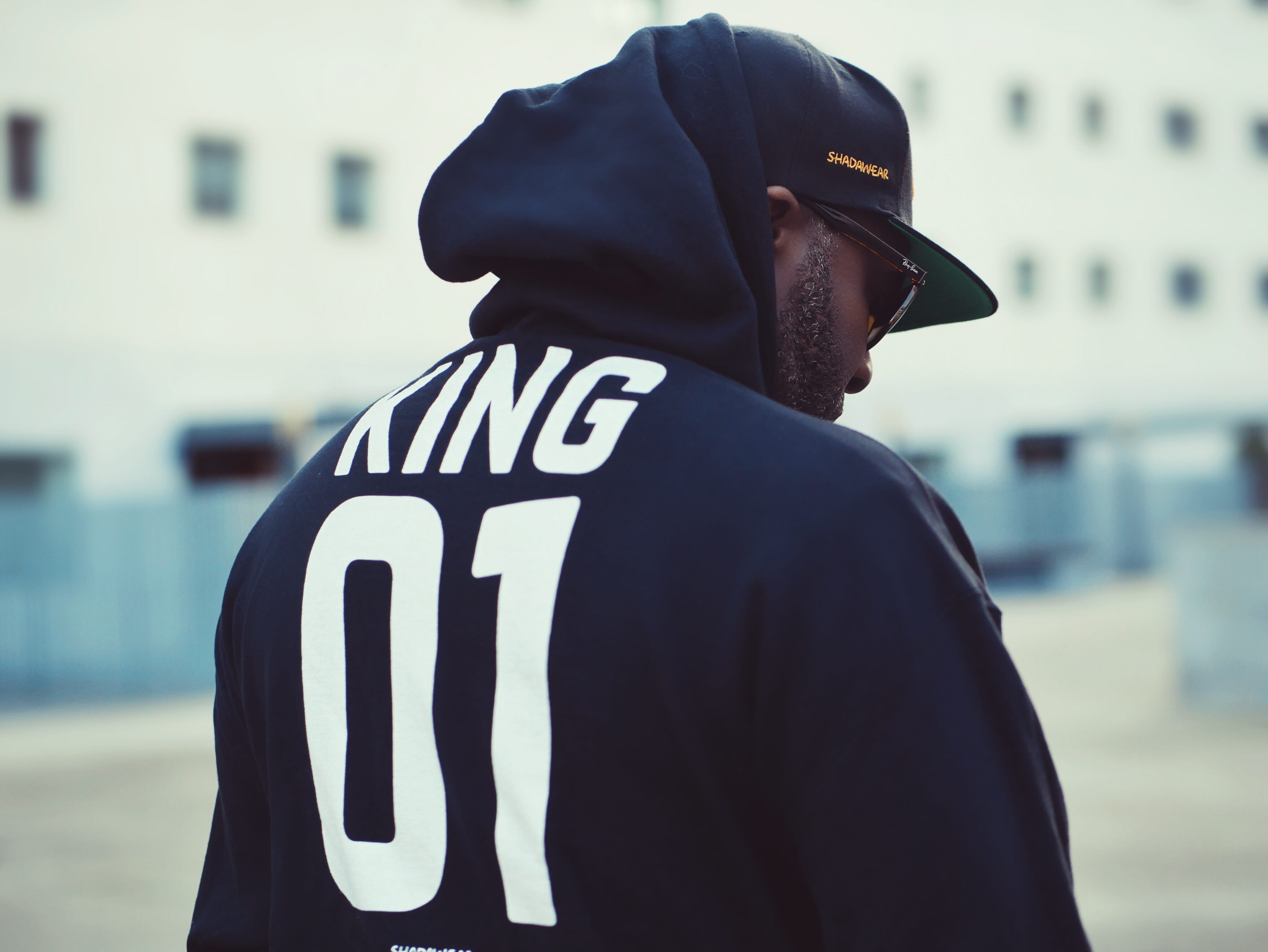 casual clothing, blur, standing, building, cap, wearing, city, one person, safety, hoodie, printed, waist up, lifestyles, day, outdoors, brim, king, uniform, HD Wallpaper