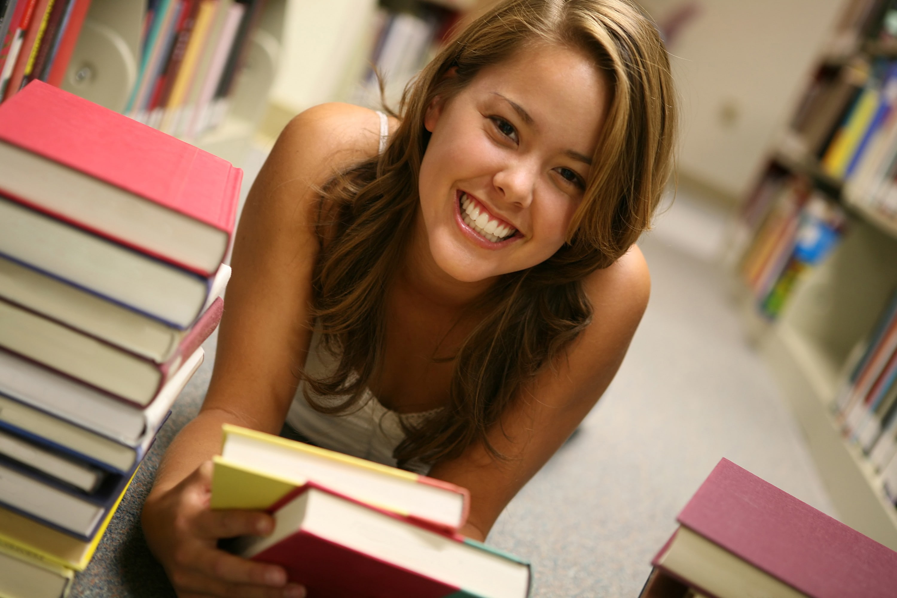 looking at viewer, long hair, 2K, tank top, lying down, women, model, library, books, shelves, bare shoulders, smiling, brunette, on the floor, HD Wallpaper