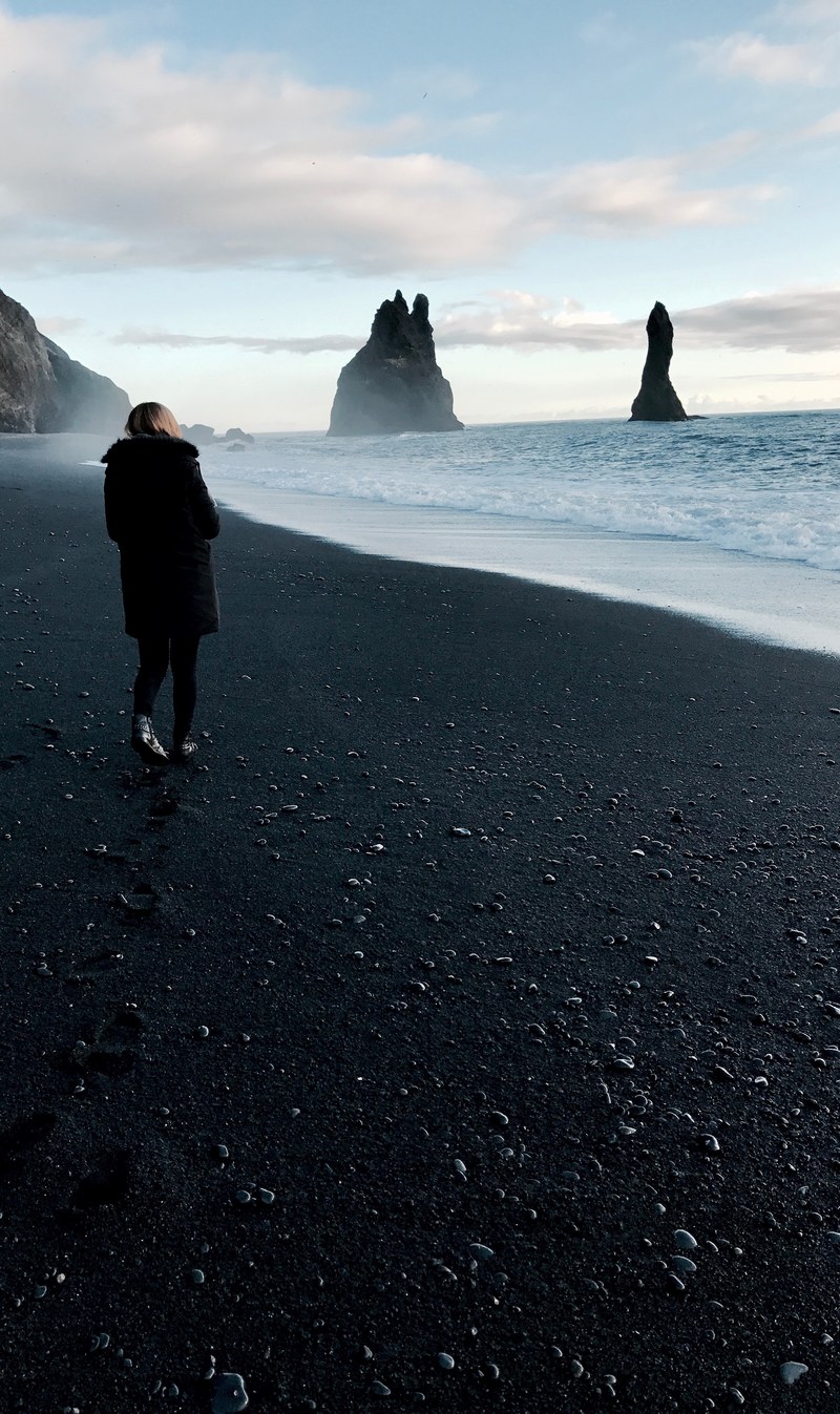 woman in black coat standing on a black sand beach looking at the ocean in iceland, icelandic black sand beach 4k