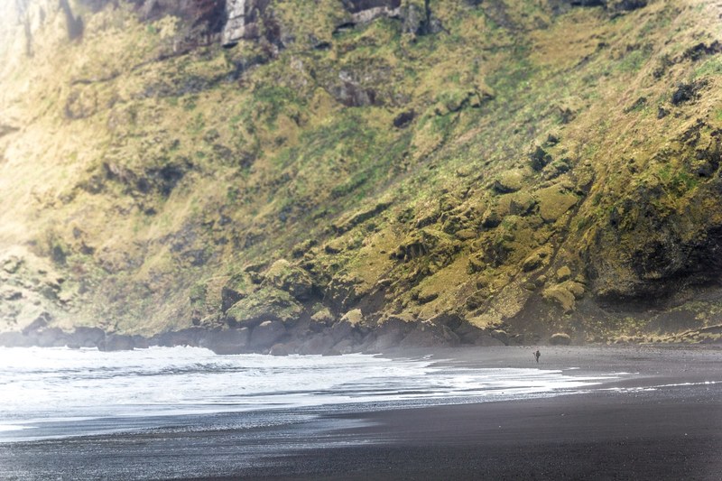 a shingle beach under a mossy cliff with a silhouette of a person in the distance, shingle beach by a cliff 4k