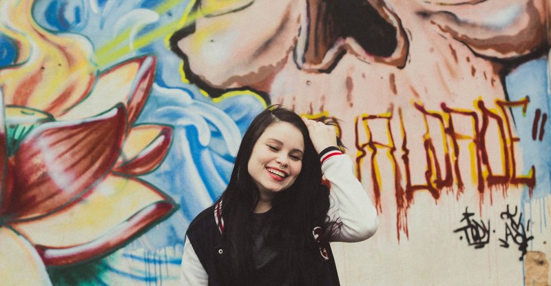 young smiling hipster girl in baseball jacket in front of colorful graffiti mural, laughing girl in front of graffiti 4k