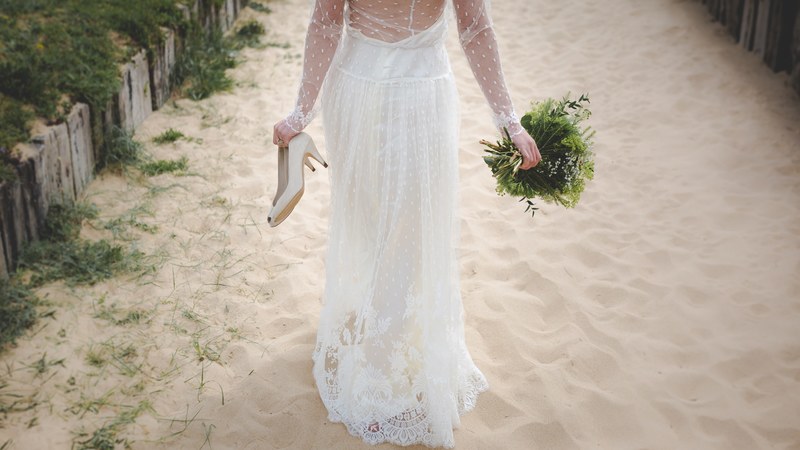 bride holds shoes and bouquet while walking in sands of sainte marie de r, bride walking in sand 4k